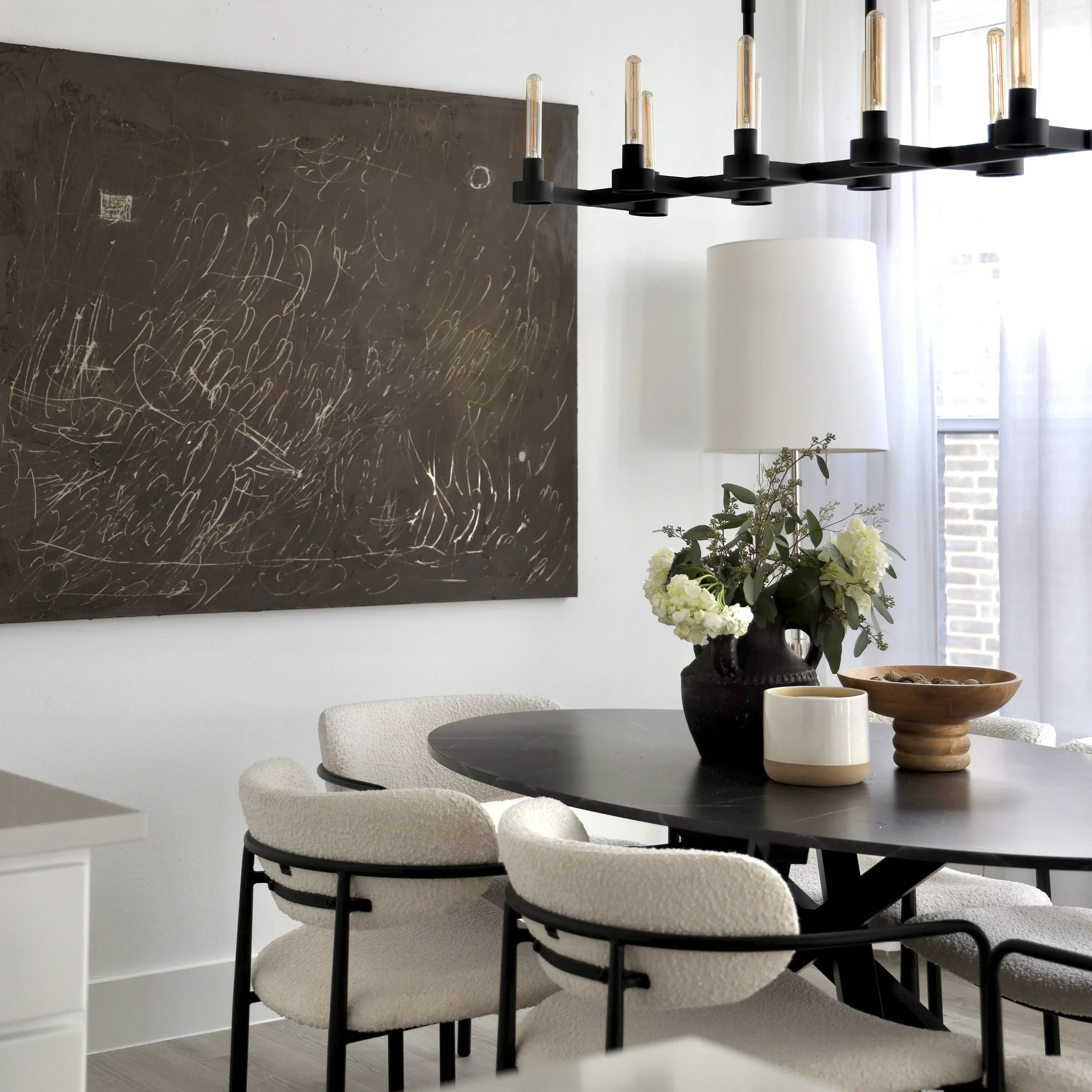 Modern dining room with black round table, white upholstered chairs, a large white lamp, black vase with white flowers, wooden bowl, and black chalkboard wall with scribbles.