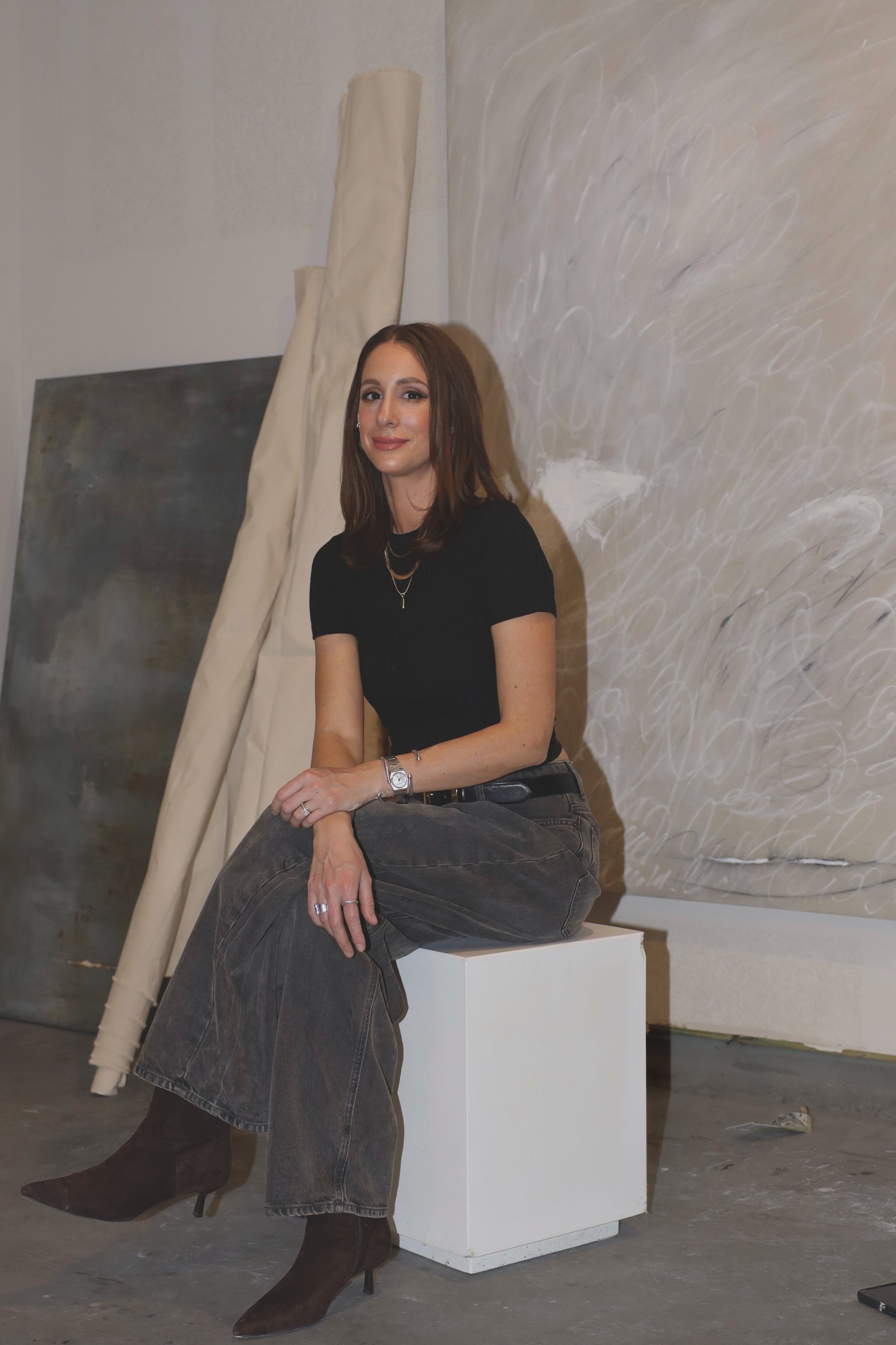 A woman with brown hair and a black top sitting on a white box in an art studio, with canvases and artwork in the background.