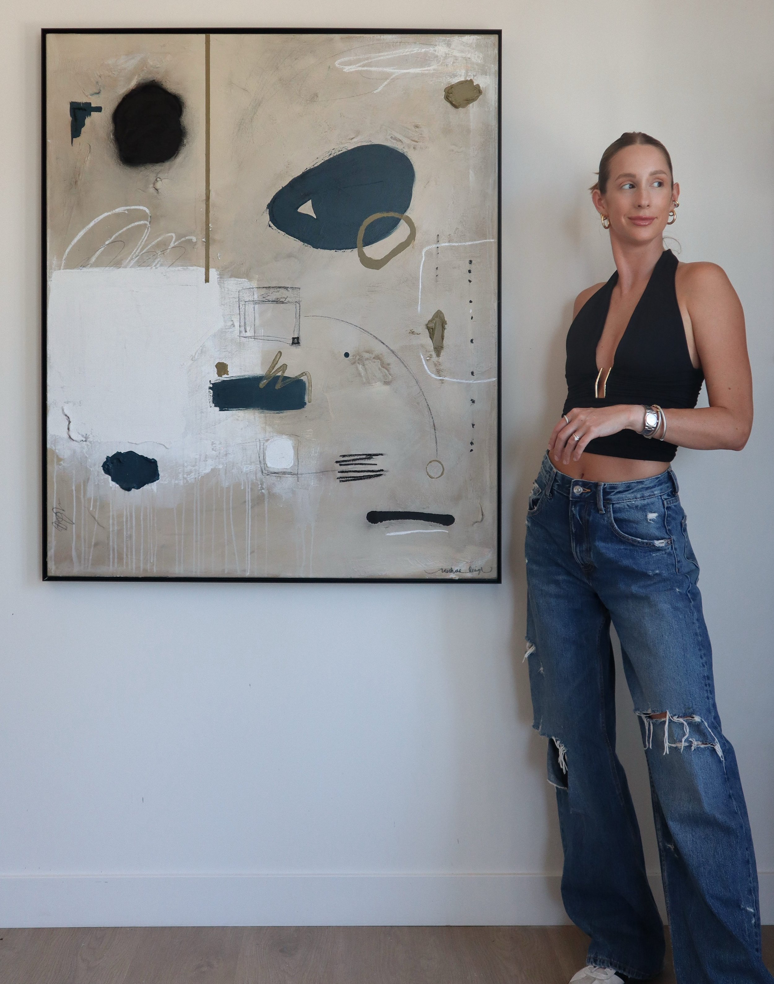 A woman in a black crop top and ripped jeans standing next to an abstract painting on a white wall.