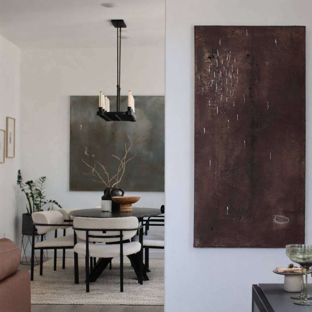 A modern dining area with a round table, white cushioned chairs with black metal frames, and a black chandelier hanging from the ceiling. There are framed pictures on the wall, a large greenish artwork, and a brown decorative bowl with branches on th