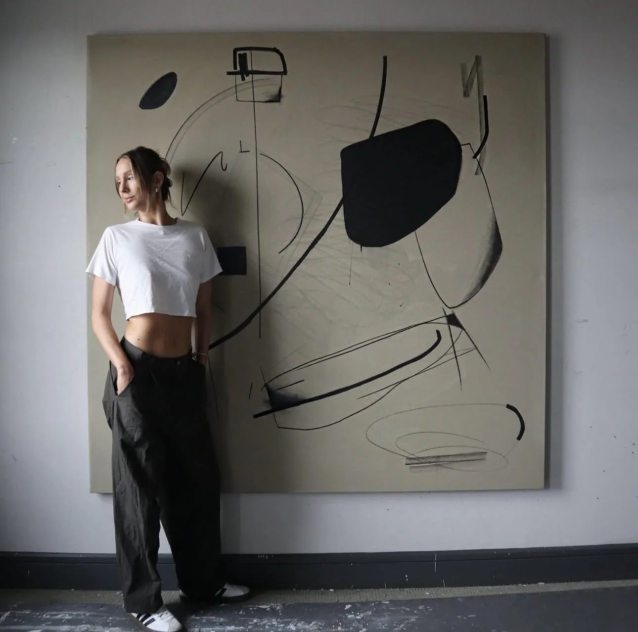 A woman in a white crop top and dark pants standing against an abstract black and white painting on the wall.