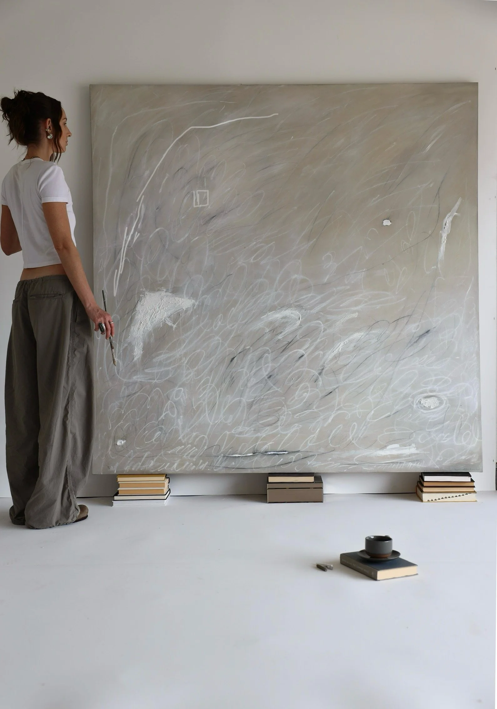 An artist stands in front of a large abstract painting with swirling white and gray lines in a gallery space, with books placed underneath the canvas.