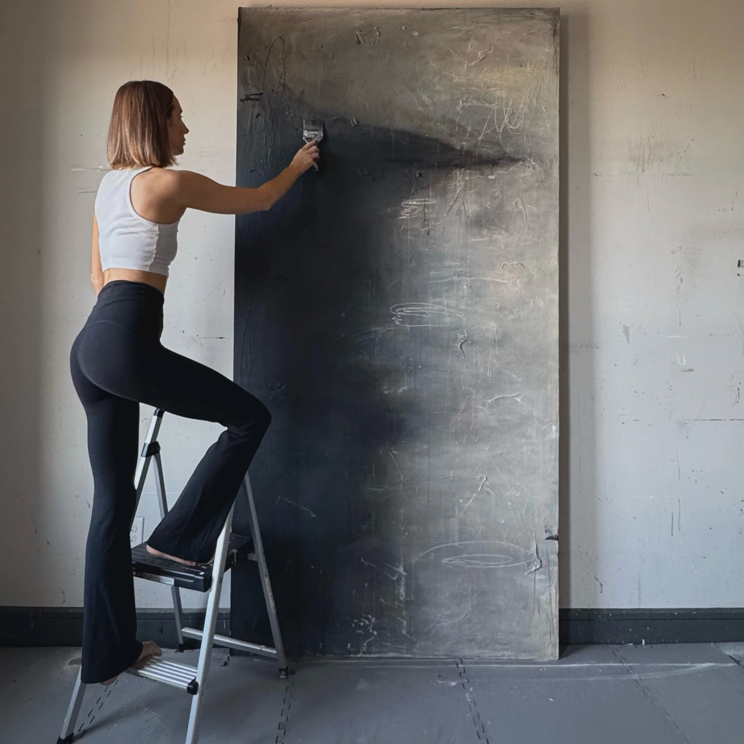 A woman standing on a step ladder, cleaning a large, dark chalkboard or writing surface with a cloth.