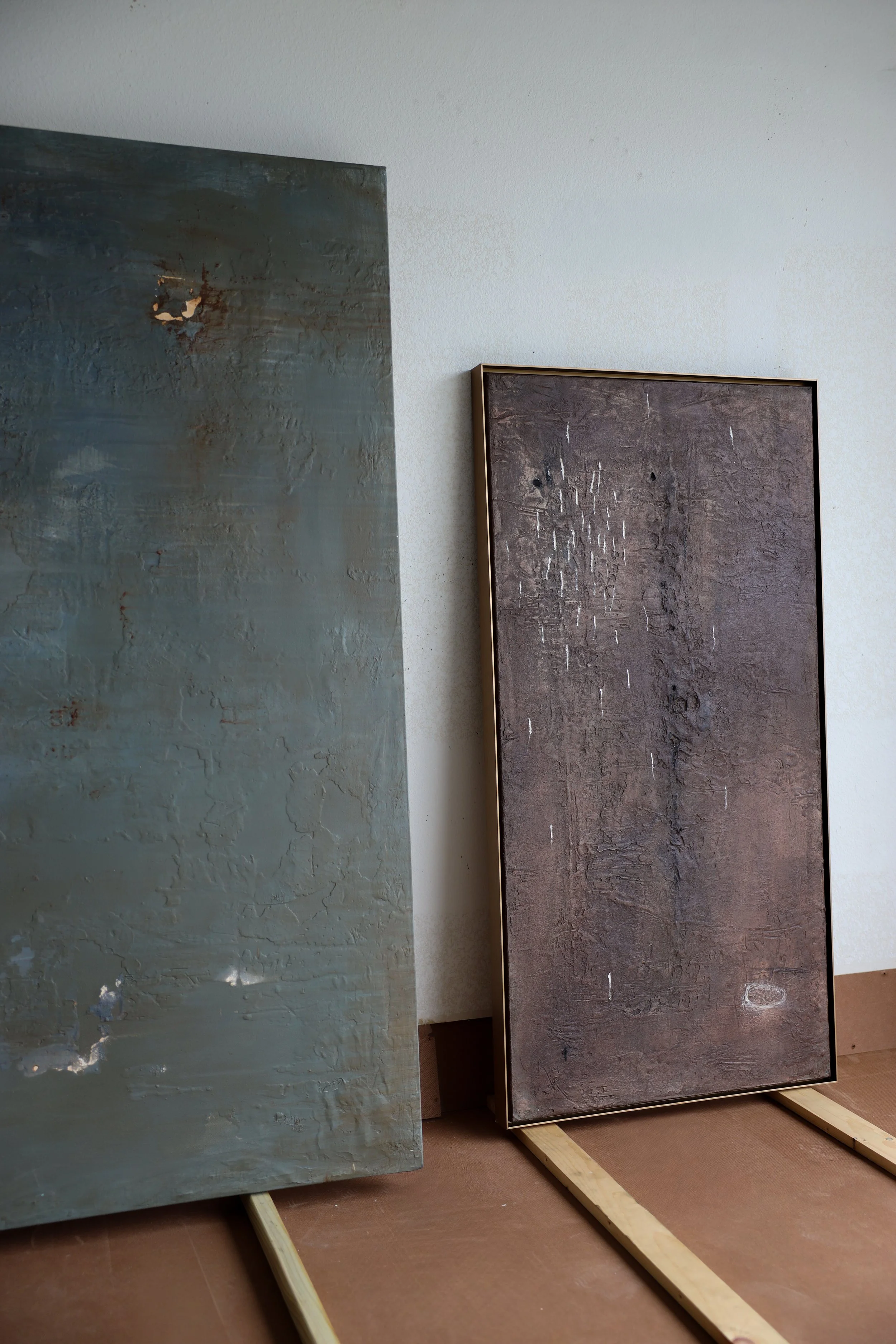 Two large canvases with textured surfaces, leaning against a white wall on a wooden floor.