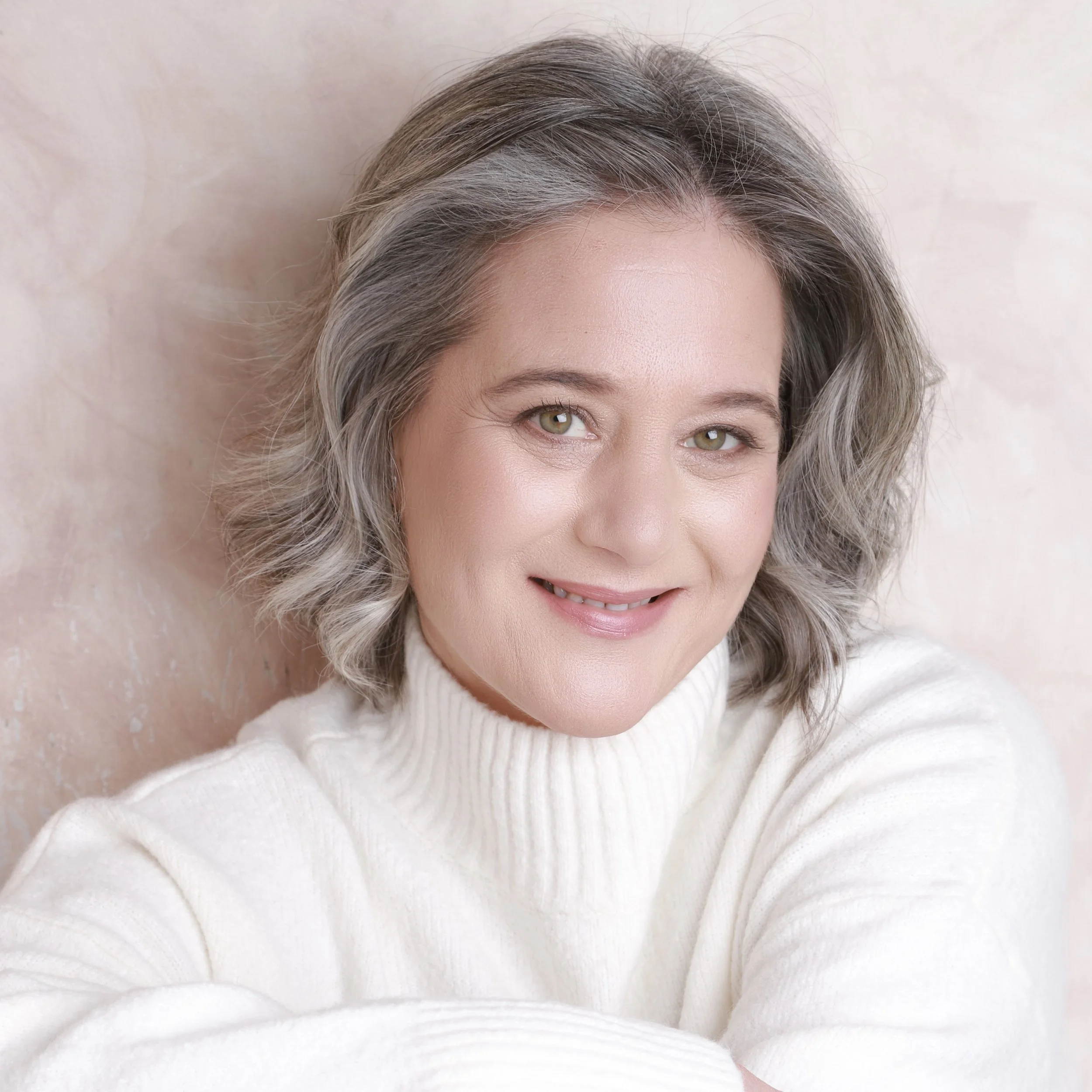 A woman with short gray hair and green eyes, smiling, wearing a white turtleneck sweater, against a soft pink background.