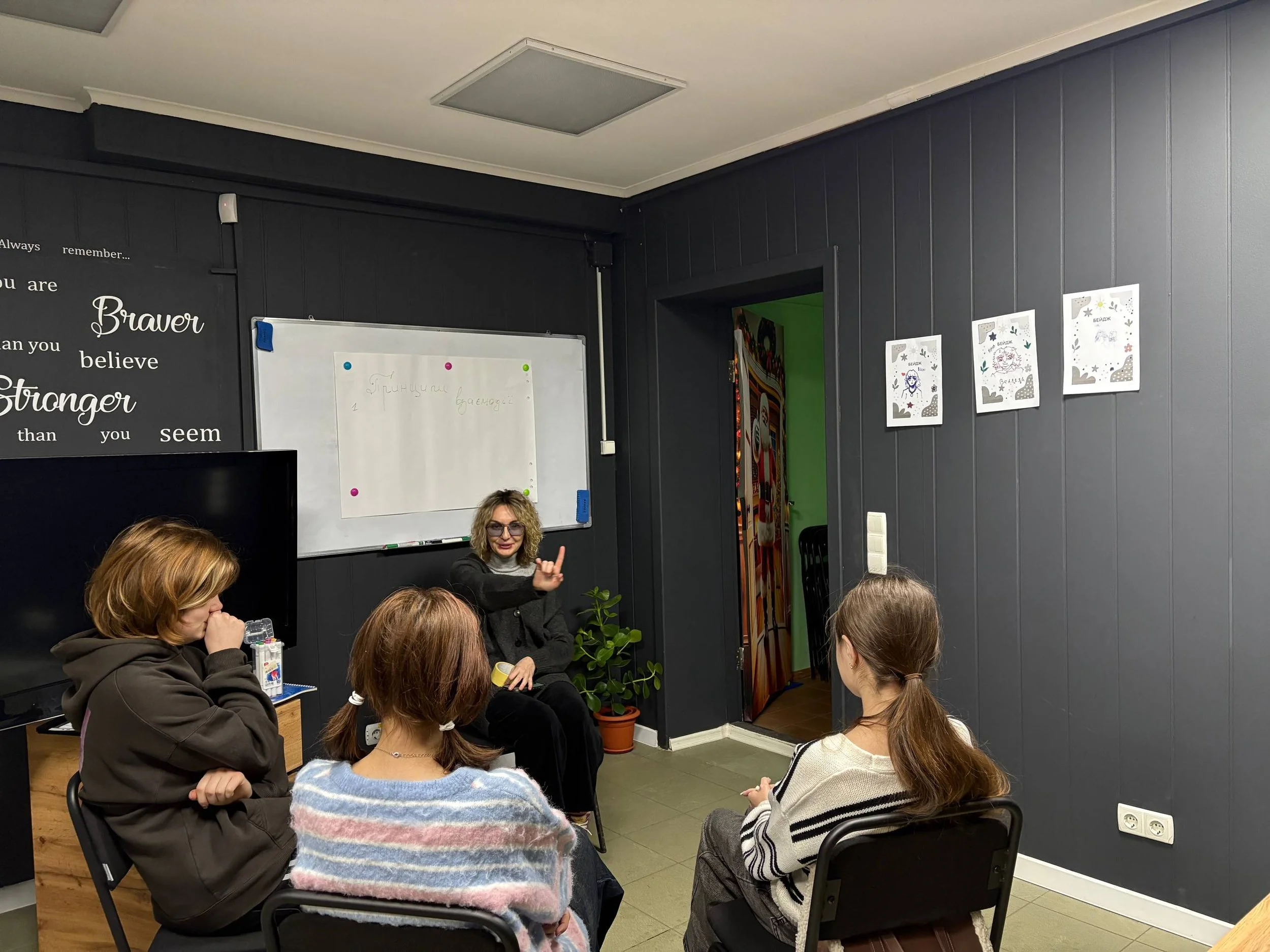 A woman with blonde hair, glasses, and a dark outfit is sitting on a chair and speaking to a group of four women seated in front of her in a room with dark gray walls. The room has a whiteboard with some writing, decorative posters on the wall, and a potted plant near the woman.