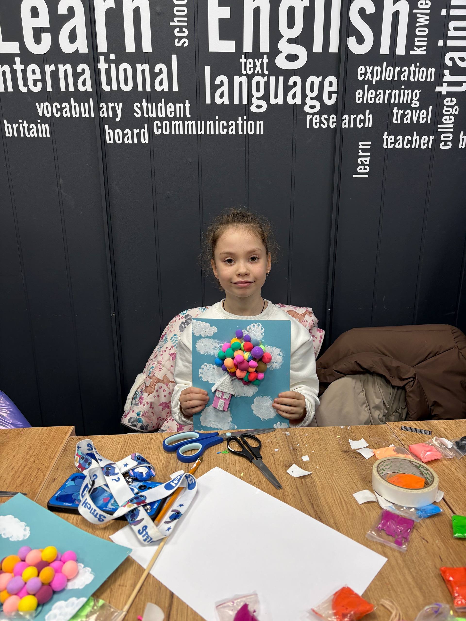 A young girl at a craft table holding a colorful art project with pompoms on a blue background.