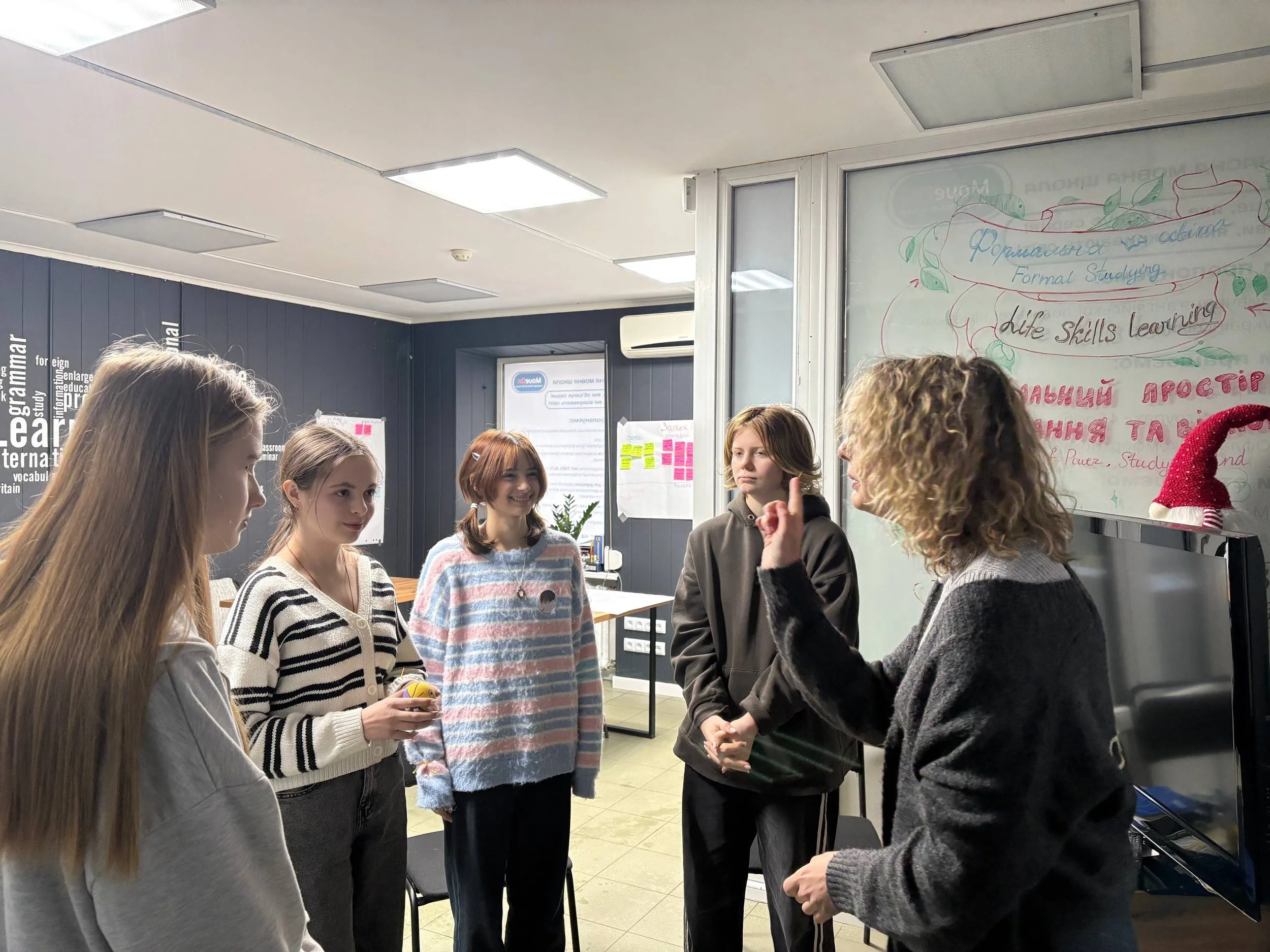 Group of five young women standing in a circle, participating in a discussion in a classroom or office space with a whiteboard and posters on the walls.