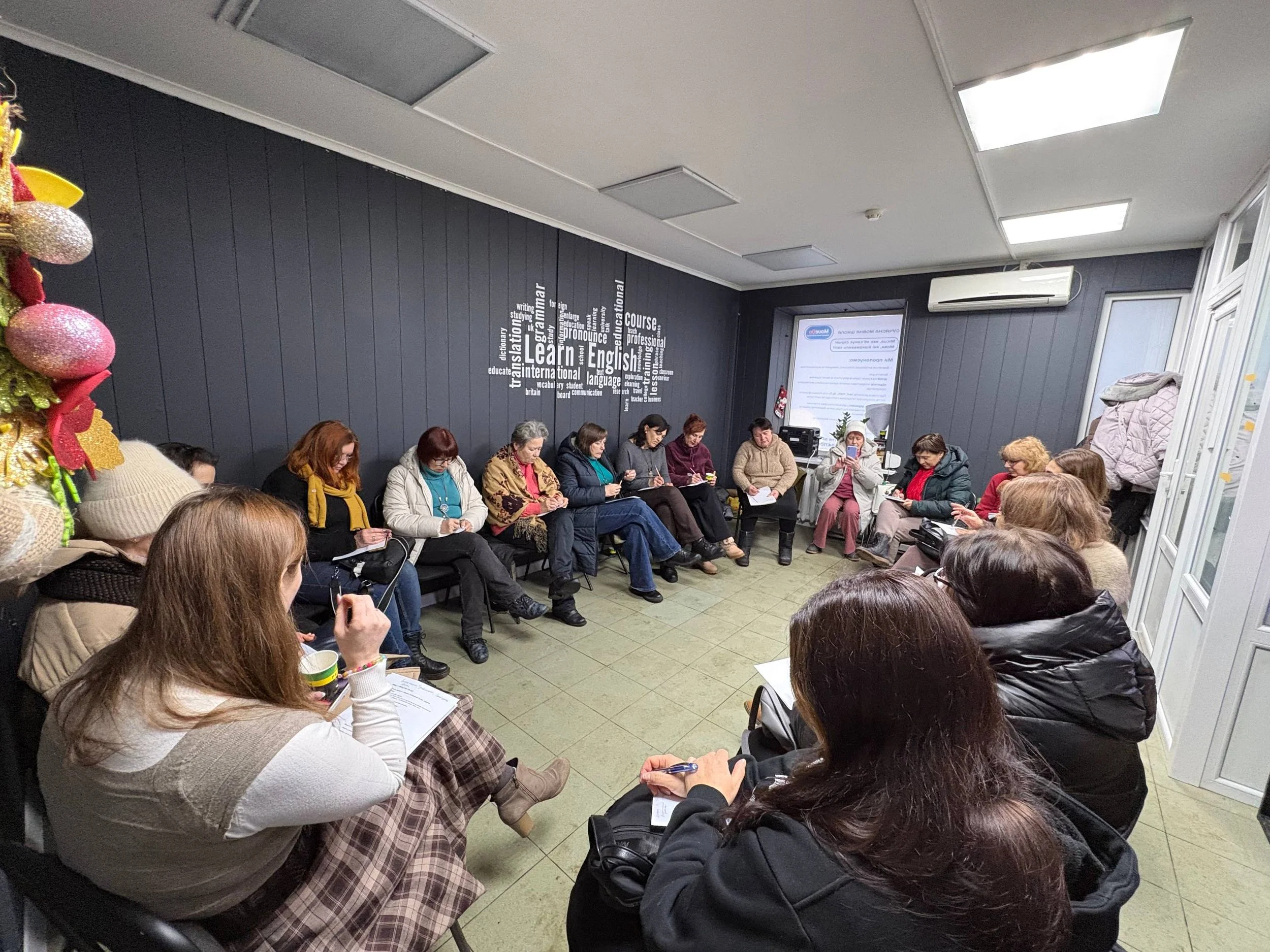 A group of people seated in a circle in a classroom or seminar room, taking notes and listening to a speaker or instructor at the front. There is a window and a large screen with information on it.