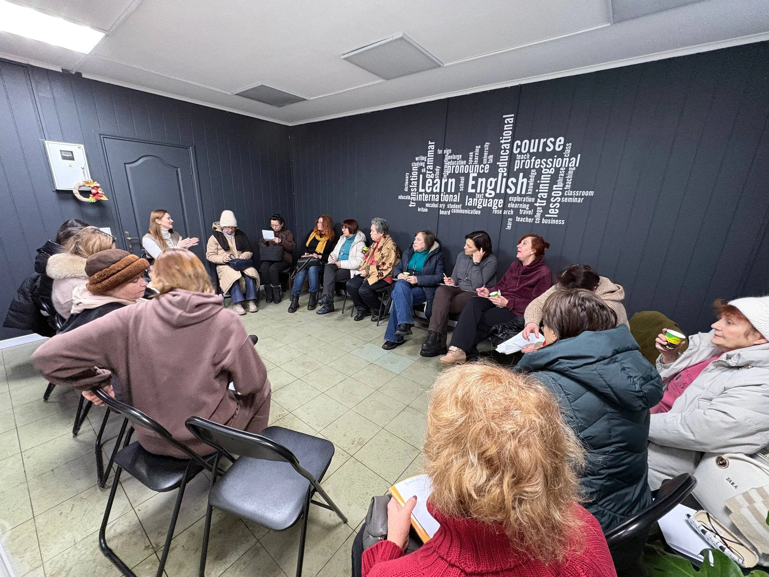 A group of people sitting in a U-shaped arrangement in a room with dark blue walls, during a discussion or class, with some holding notebooks or cups, against a wall with a large word cloud related to learning English.