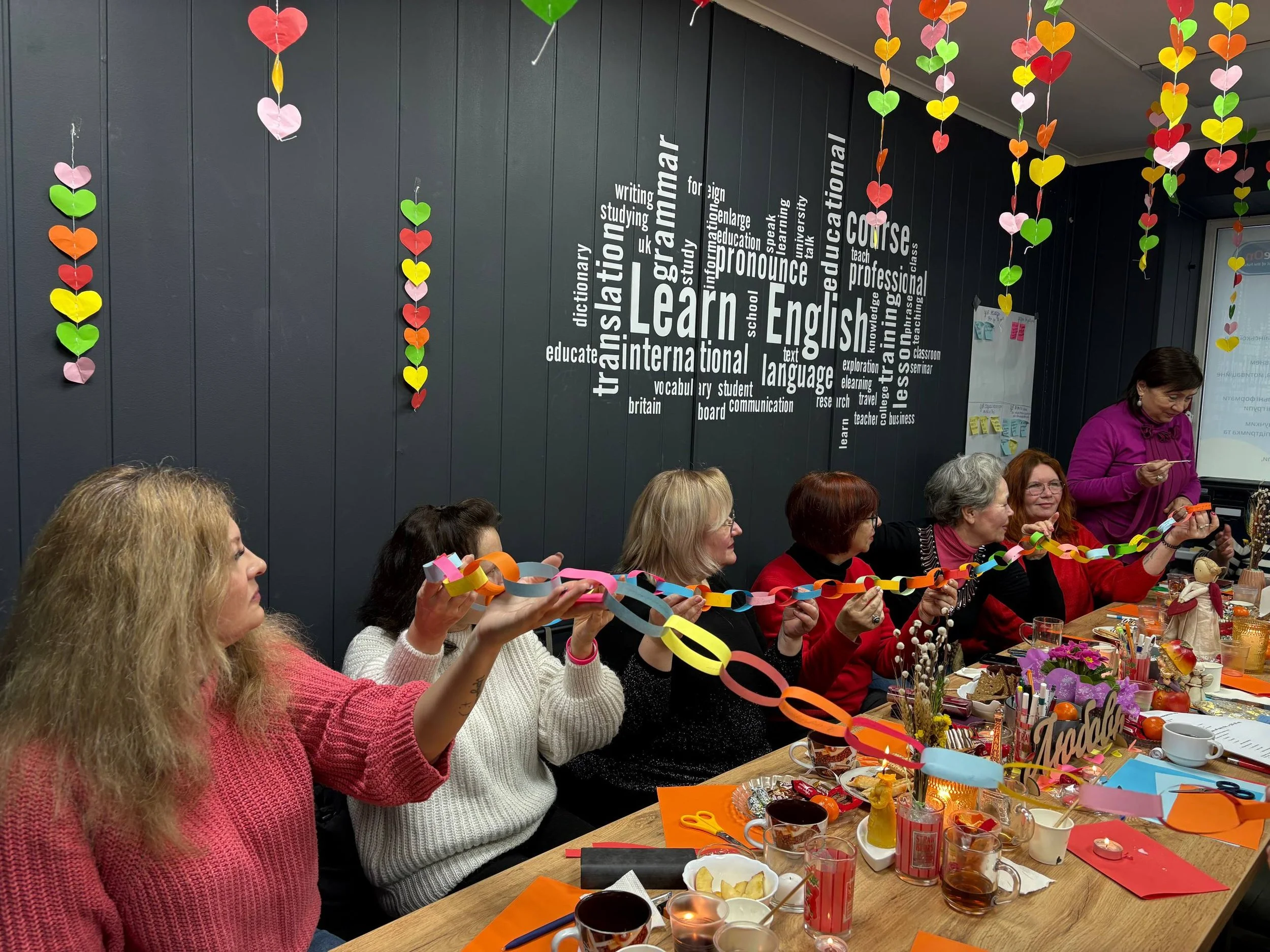 A group of women sitting around a table decorated for a celebration, holding a colorful paper chain. The room has hanging paper hearts and a wall with words related to learning English.