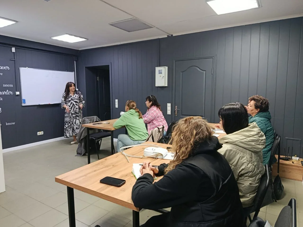 A woman standing at the front of a classroom or meeting room speaking to five women seated at wooden tables. The room has dark gray wooden walls and a whiteboard on the wall.