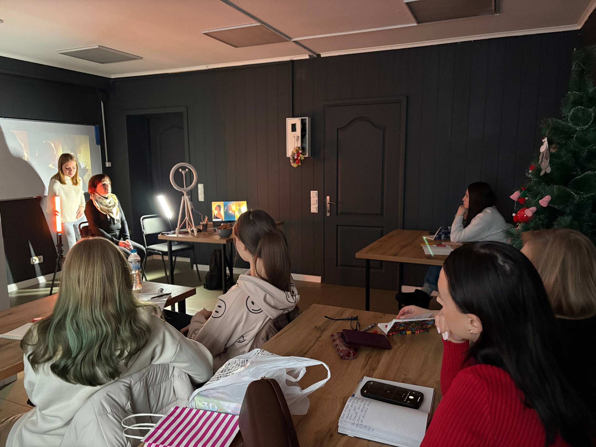 A group of women seated around a wooden table watching two women perform a skit or presentation in a dark room decorated with a Christmas tree.