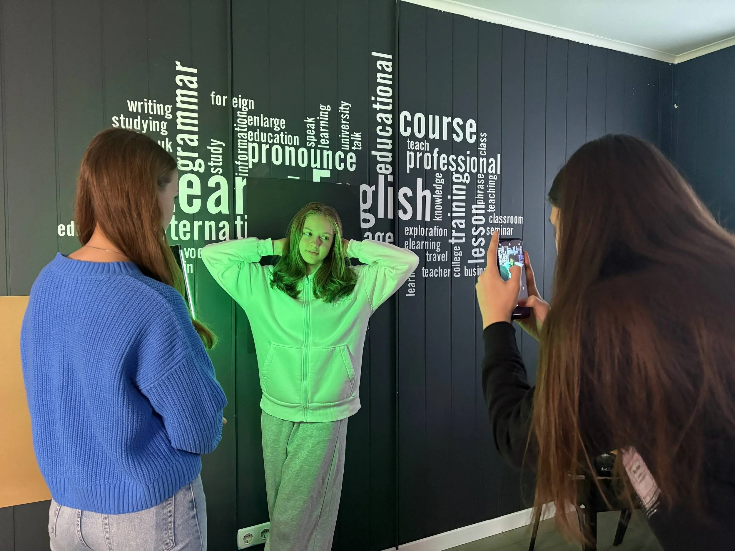 Three teenage girls with long hair, two with brown and one with blonde, are in a room with a dark wall displaying words related to language learning. One girl in a black sweatshirt is taking a picture of a girl in a light-colored hoodie with her smartphone. The girl in the hoodie is posing with her hands behind her head.