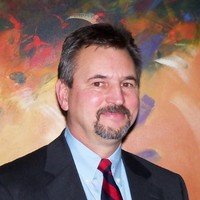 A man with short dark hair and a beard, wearing a dark suit jacket, blue shirt, and a red tie, smiling in front of a colorful abstract background.