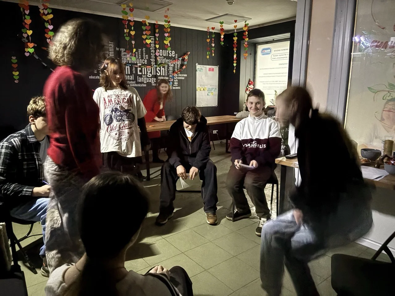 Group of children gathered in a room with black walls and decorated with colorful paper hearts hanging from the ceiling. Some children are sitting on chairs, and others are standing, engaged in conversation or activity.