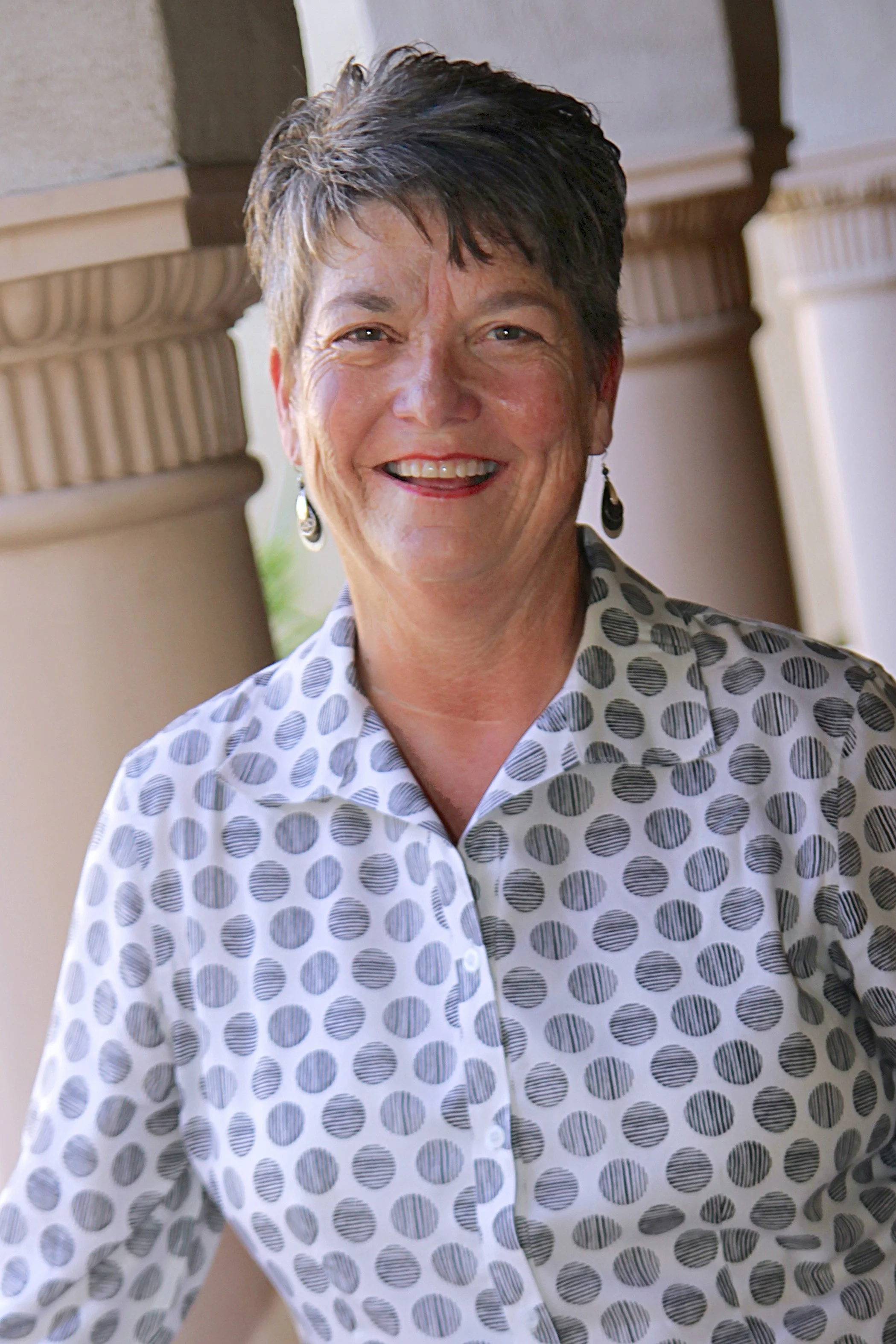 Smiling middle-aged woman with short dark hair, wearing a white blouse with black polka dots and black earrings, standing outside near columns.