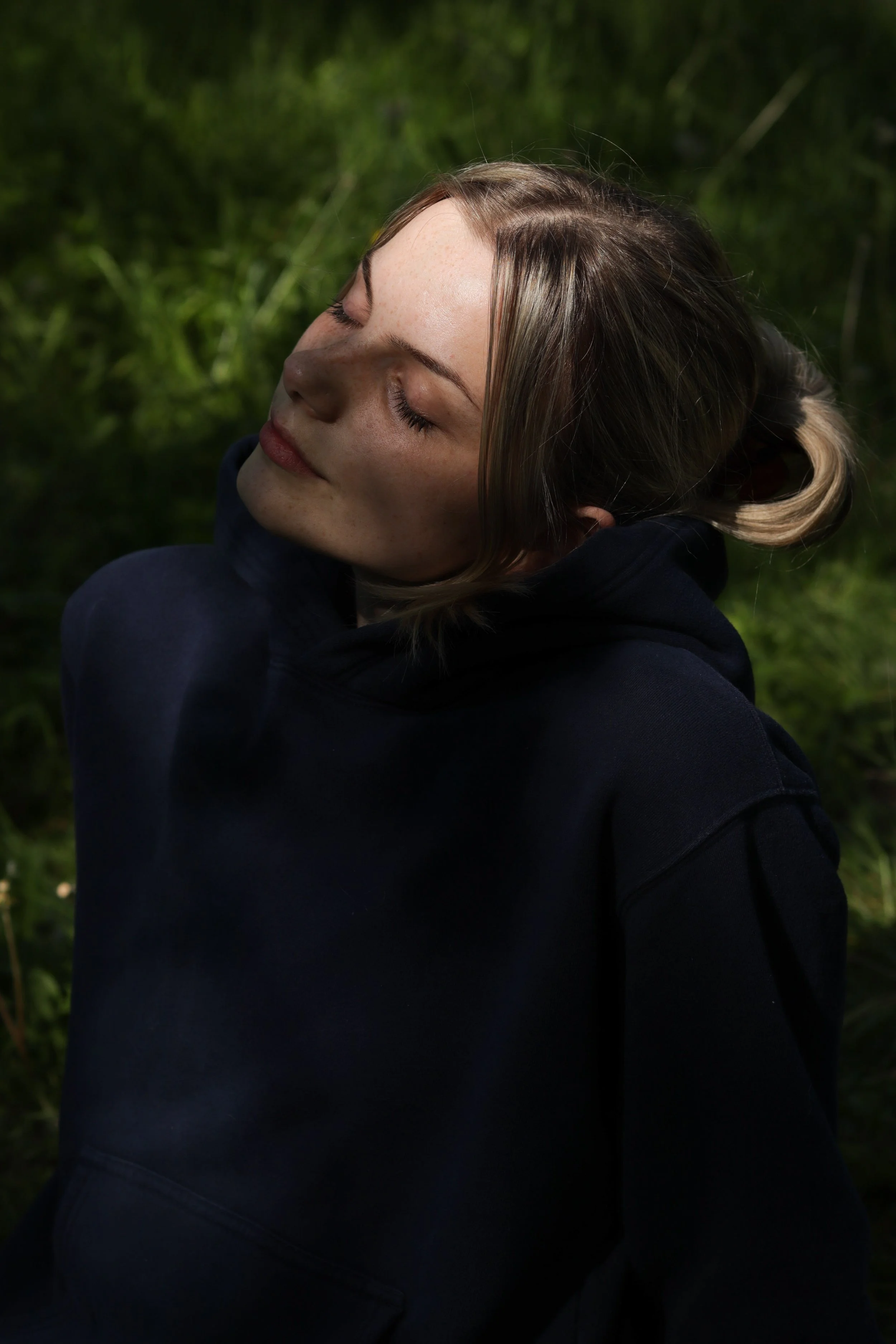 A woman with closed eyes and a serene expression, sitting outdoors in a green, natural setting, wearing a dark hoodie.