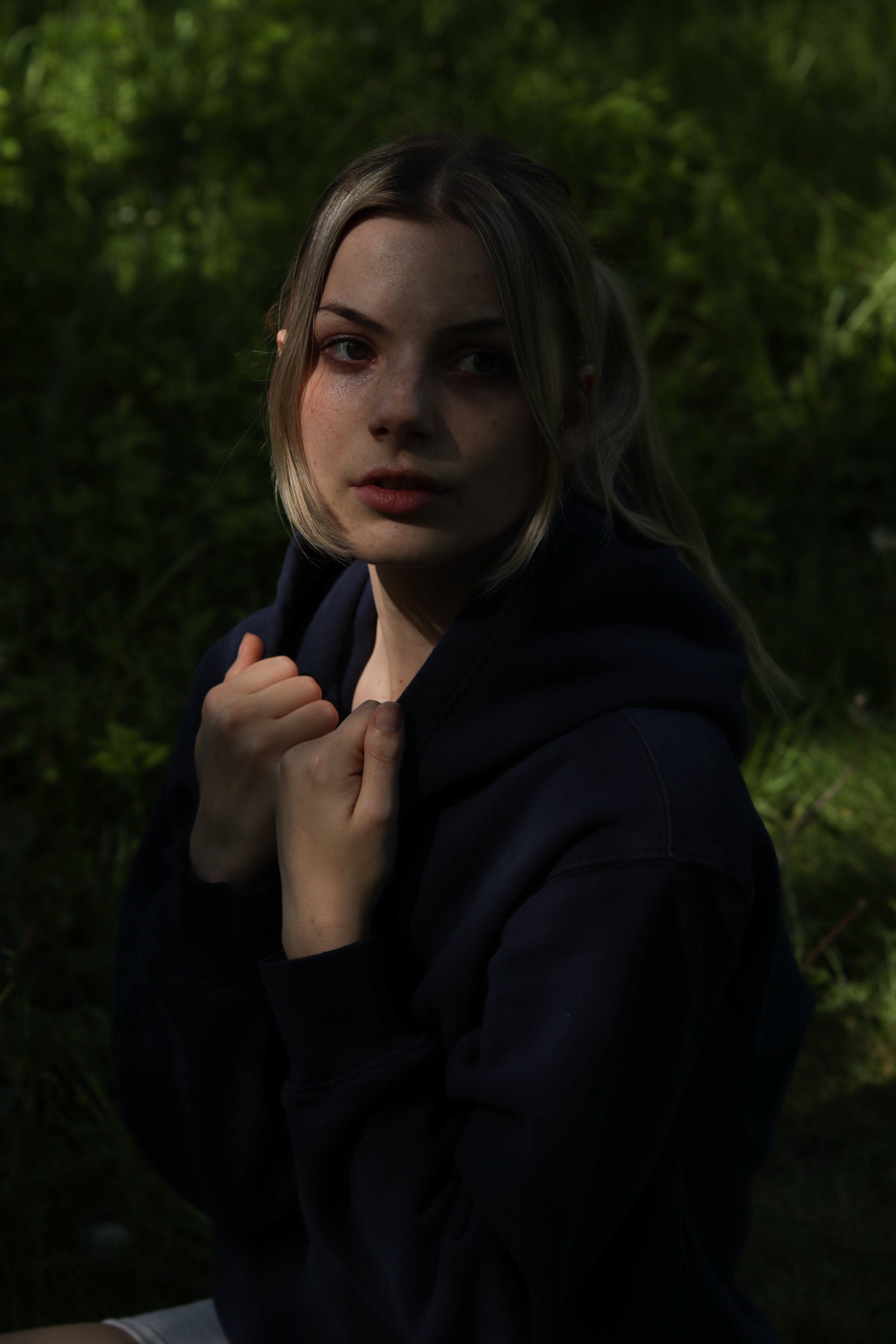 A young woman with blonde hair and freckles wearing a dark hoodie, sitting outdoors surrounded by green foliage, looking at the camera.