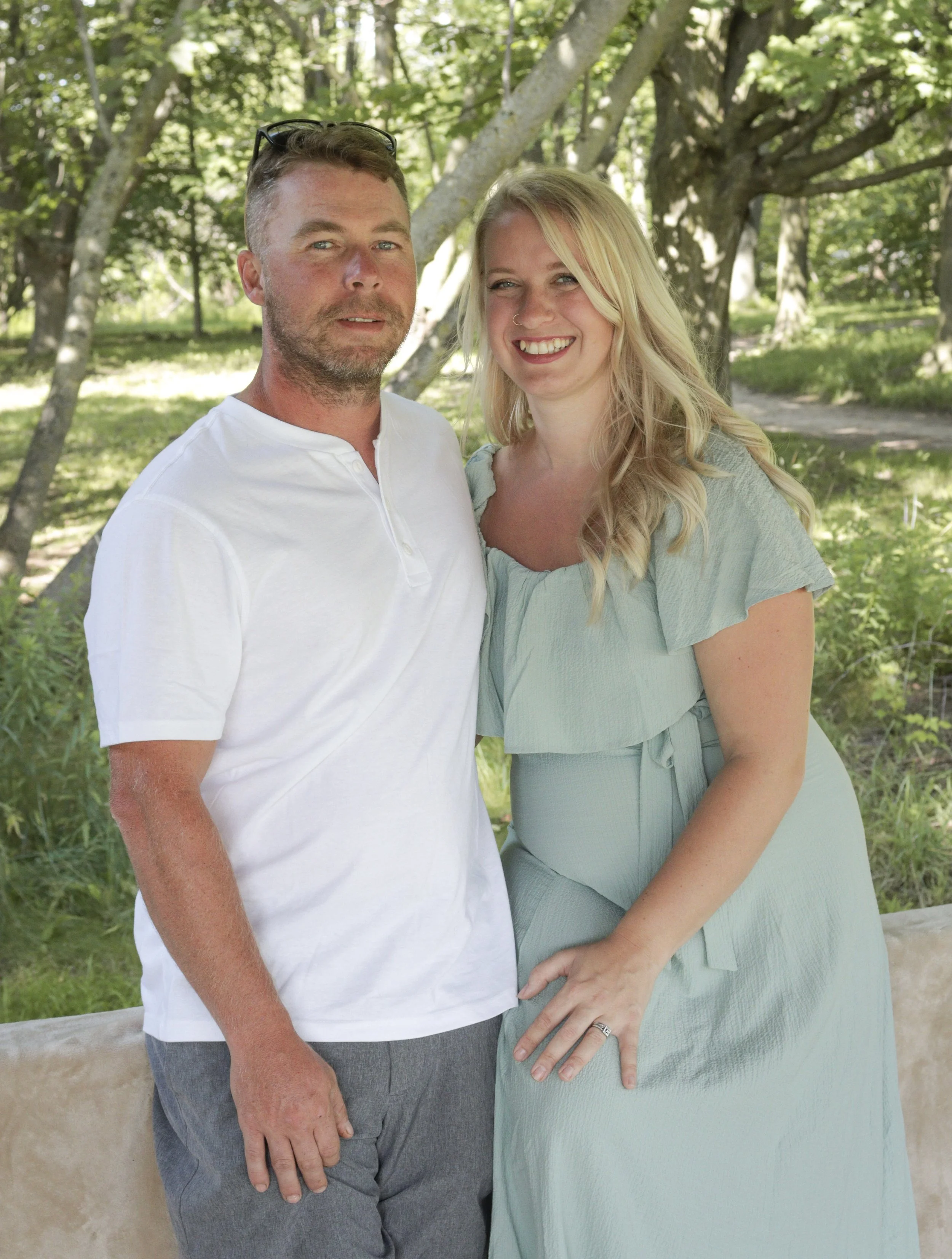 A smiling couple standing outdoors in a park-like setting with trees and greenery, the man wearing a white shirt and gray pants, and the woman in a light green dress.