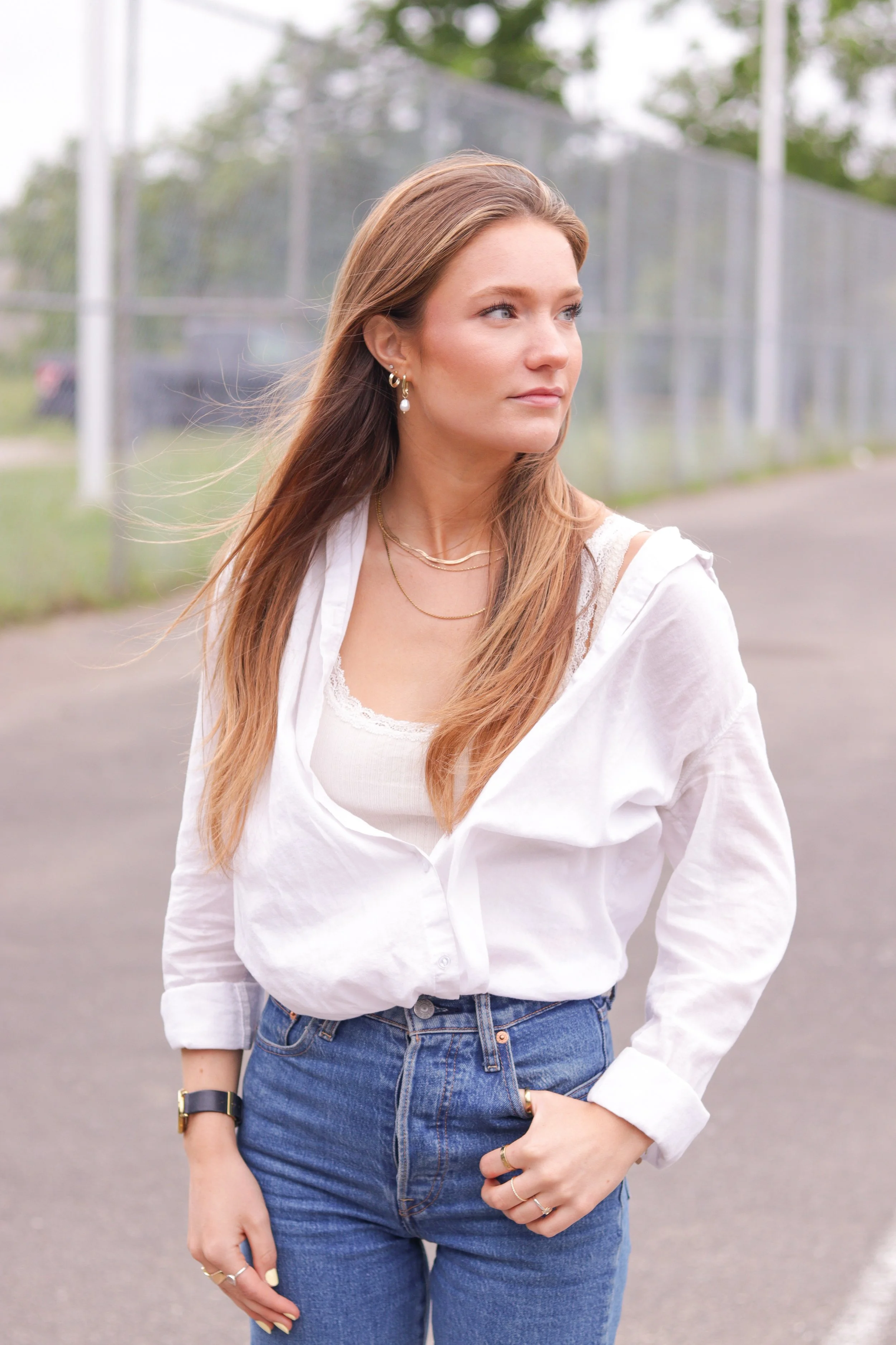A young woman with long, light brown hair, wearing a white button-up shirt over a white top with lace, and blue jeans. She has layered necklaces, hoop earrings, and a watch, standing outdoors near a chain-link fence with greenery behind her.