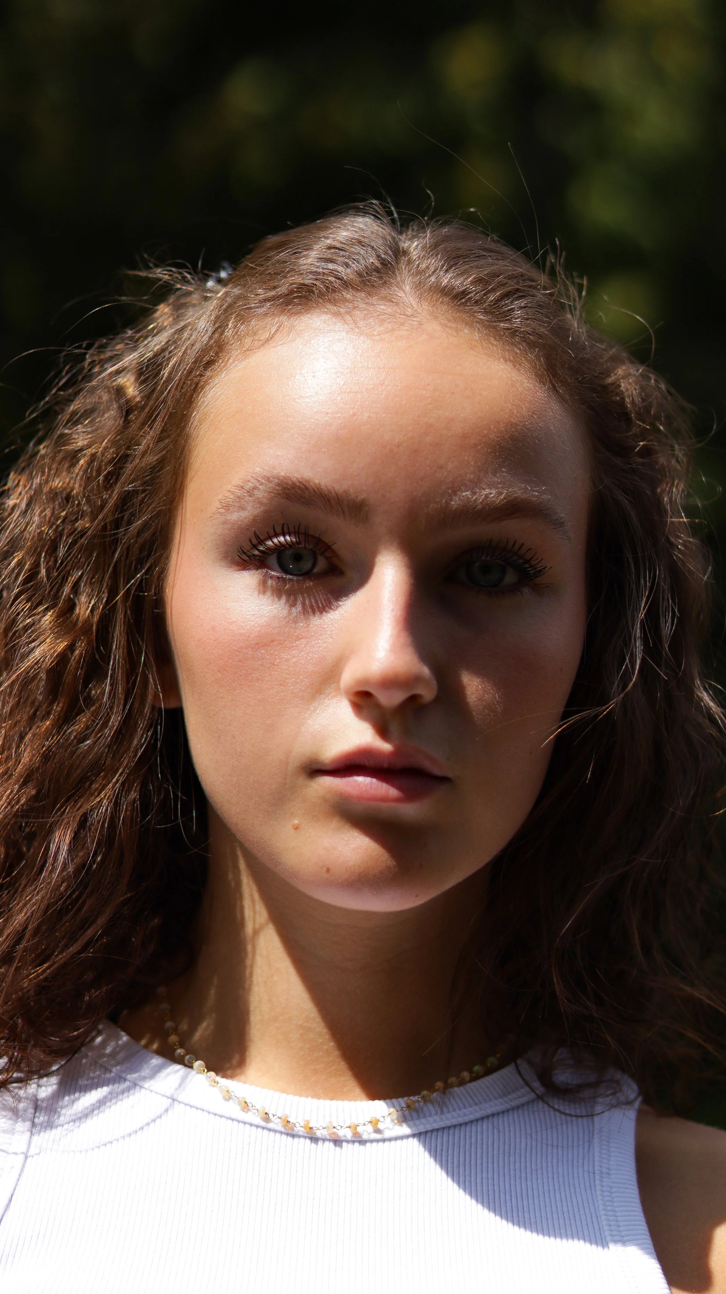 Close-up of a young woman with wavy brown hair, blue eyes, and light makeup, outdoors with a dark blurred background, wearing a white sleeveless top and a delicate beaded necklace.