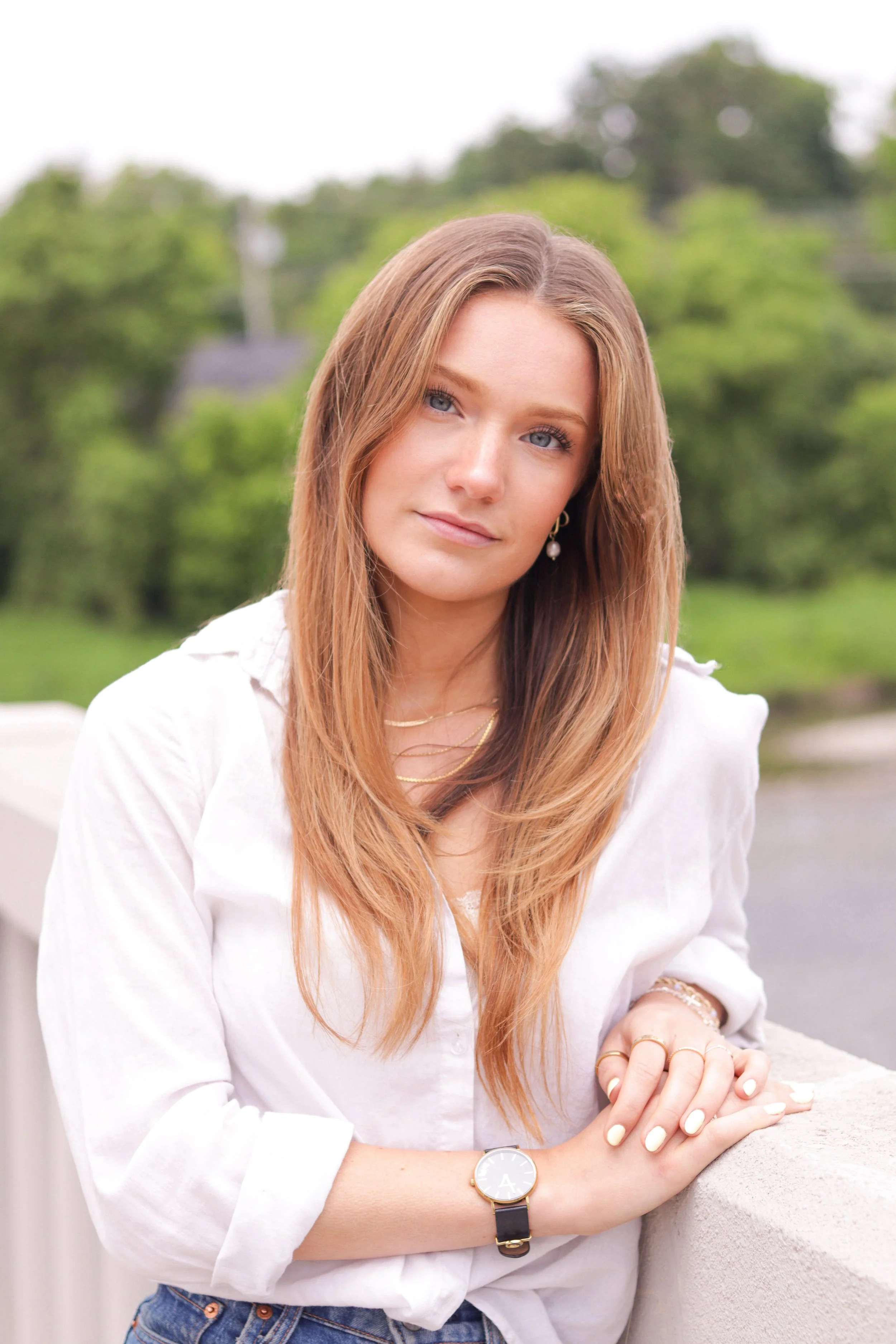 A young female model with long light brown hair and blue eyes posing outdoors, leaning on a white concrete railing, wearing a white blouse, jewelry, and a watch, with green trees and overcast sky in the background.