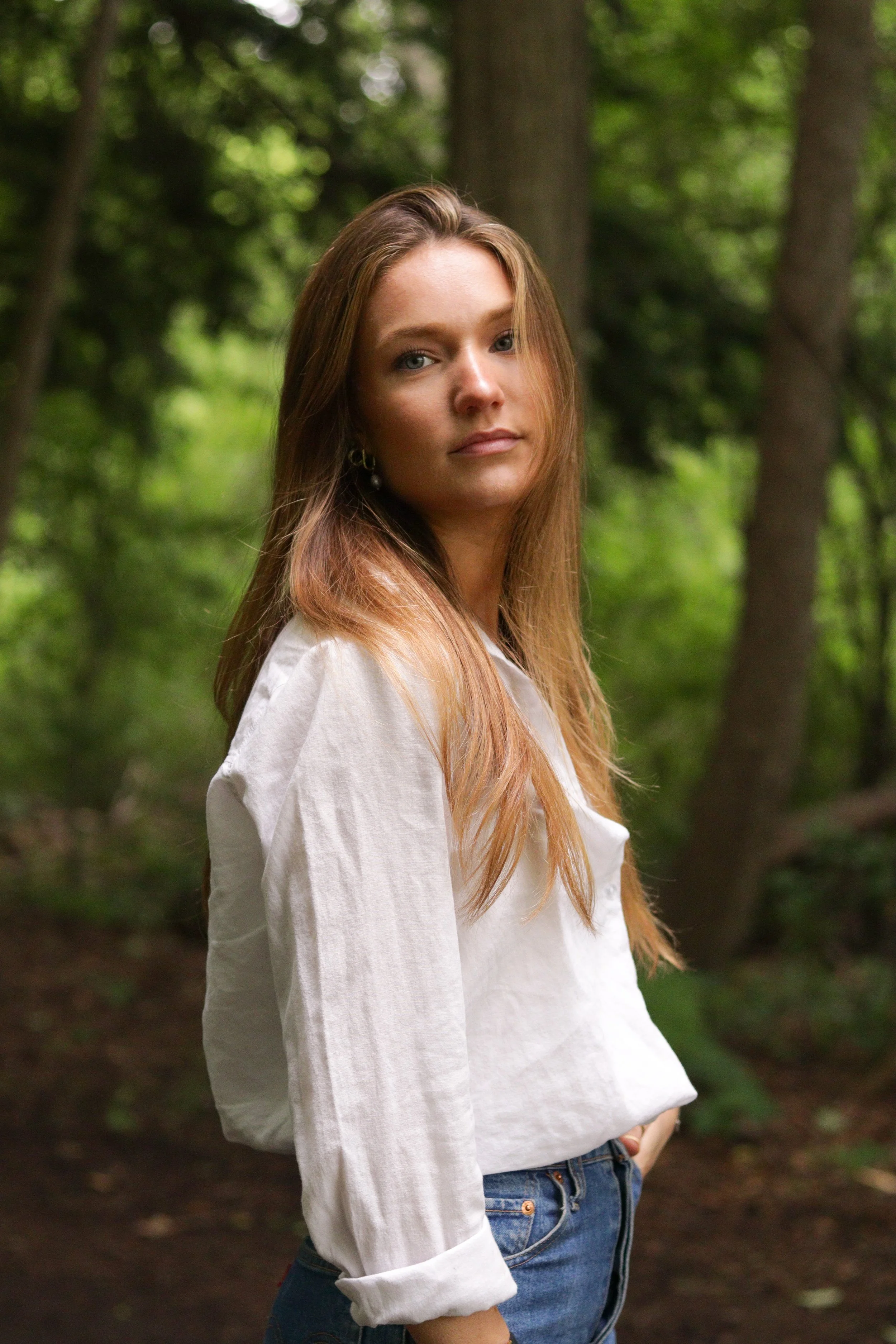 A young woman with long brown hair and blue eyes stands in a lush green forest, wearing a white long-sleeve shirt and blue jeans.