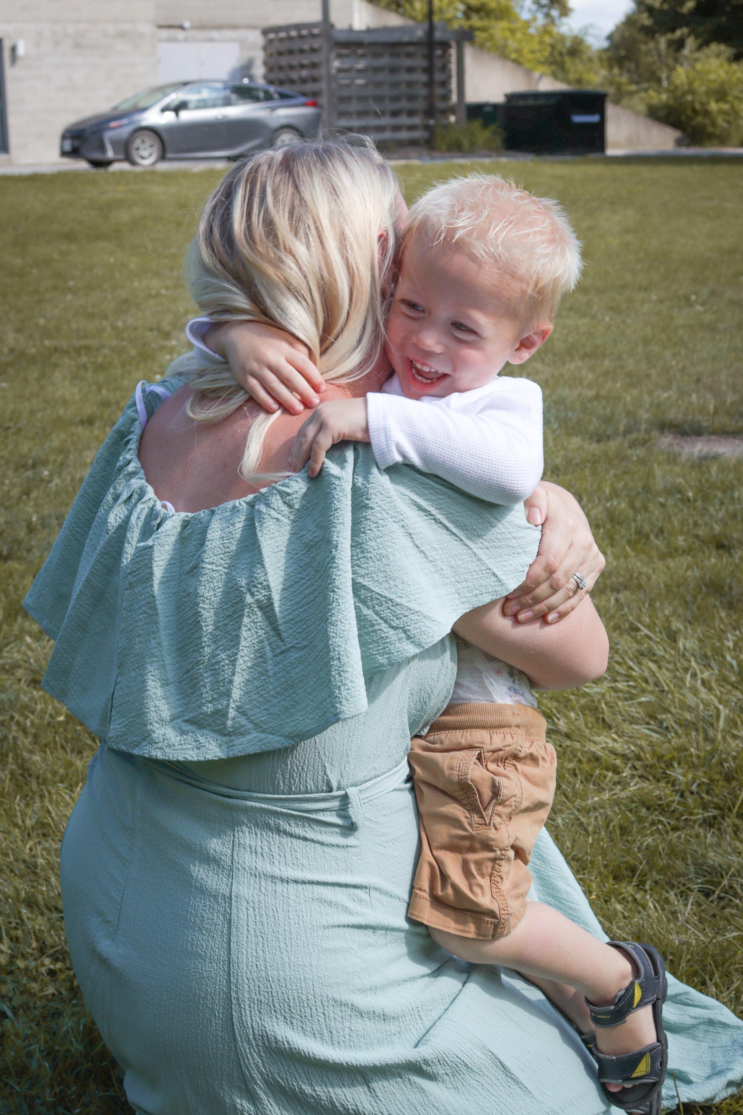 A woman with blonde hair hugging a young blonde boy in a white long-sleeve shirt outdoors on a grassy area, with a parking lot and buildings in the background.
