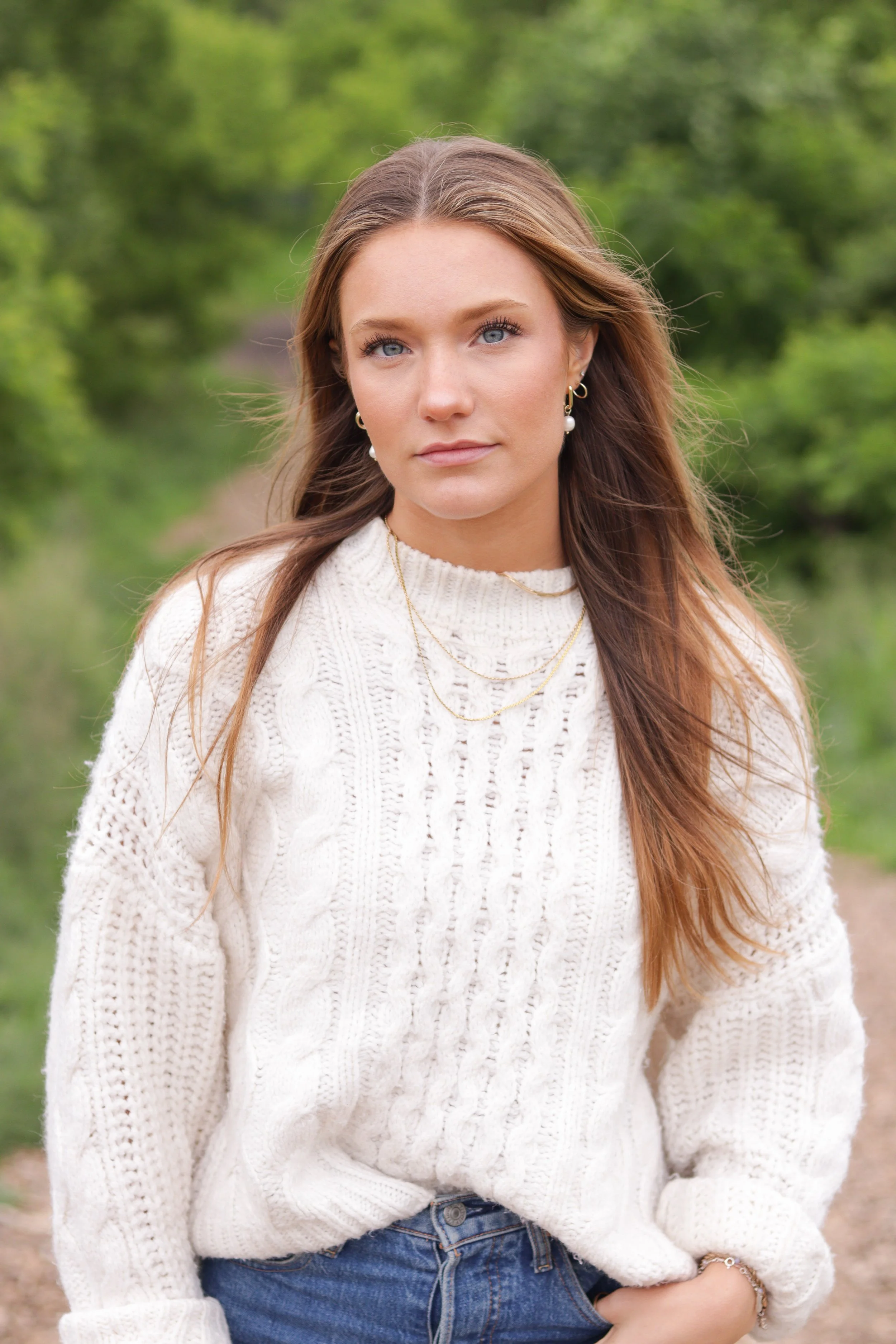 A young female model with long brown hair and blue eyes standing outdoors in a green, wooded area, wearing a white knit sweater and blue jeans.