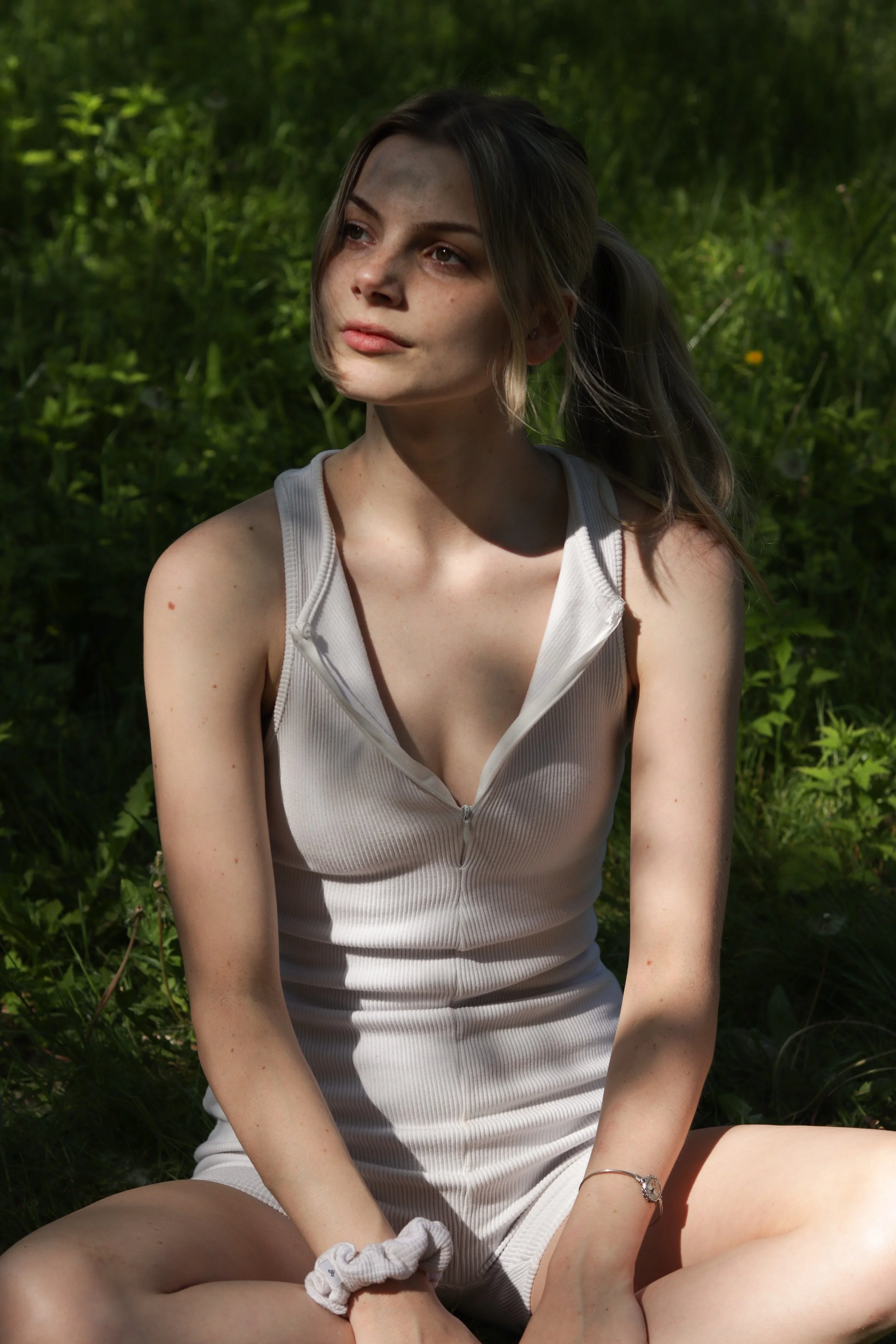 A young woman with freckles and brown hair tied in a ponytail sitting outdoors in sunlight, wearing a sleeveless beige zip-up dress, surrounded by green foliage.