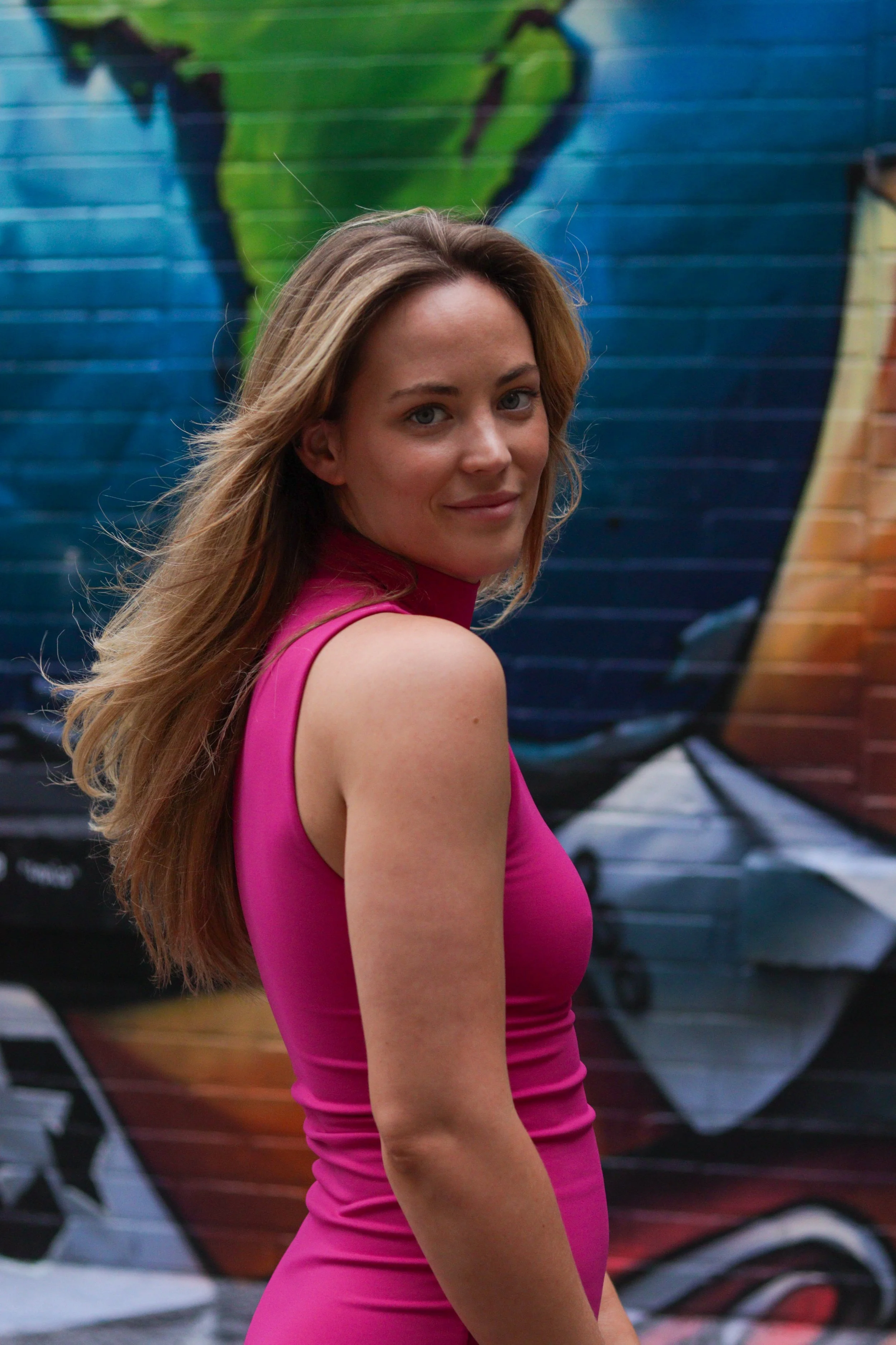 A woman with long, wavy blonde hair wearing a sleeveless, bright pink dress, standing in front of a colorful graffiti wall.