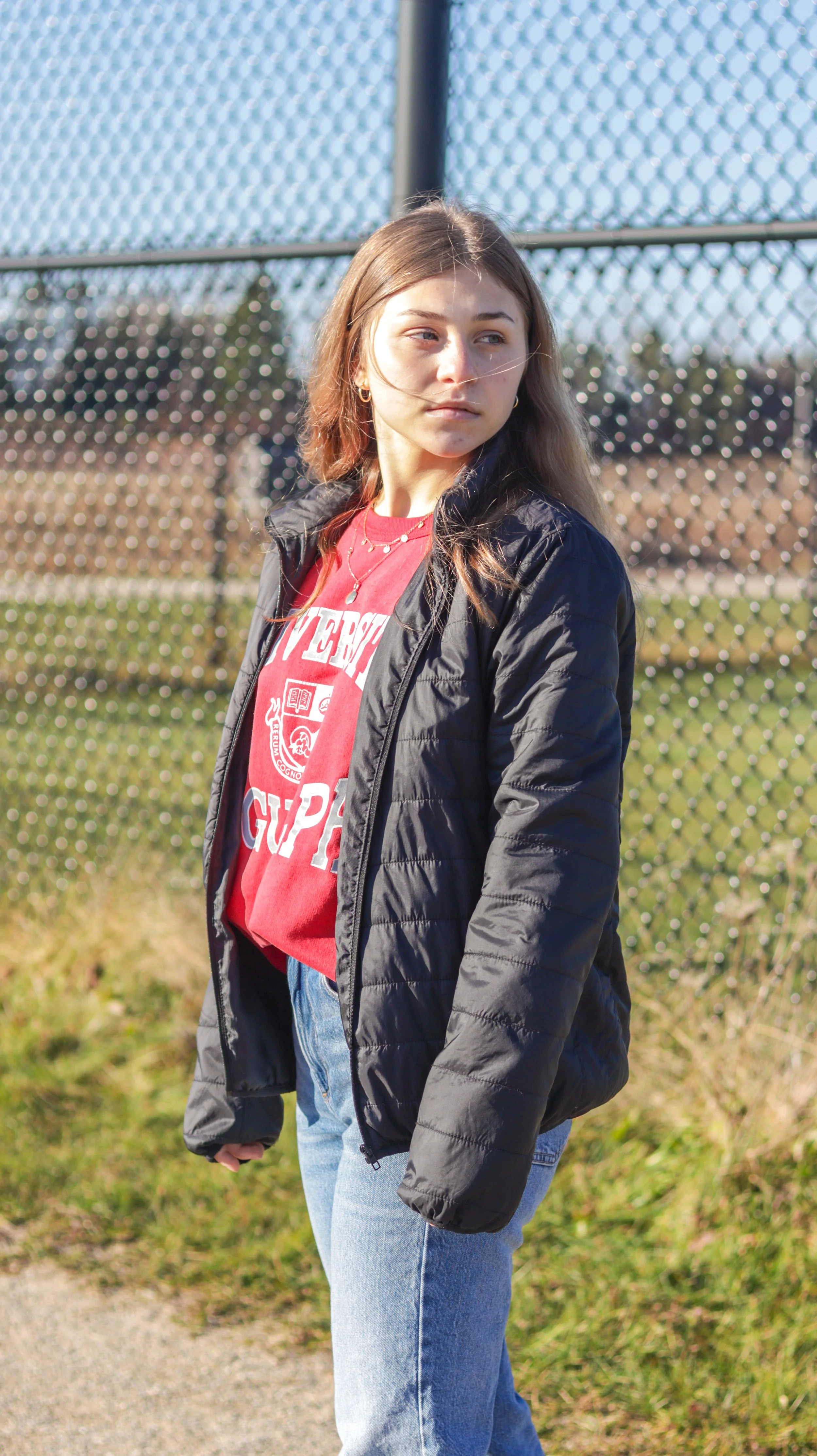 A young woman with long brown hair, wearing a red T-shirt with white text underneath a black jacket, standing outdoors near a chain-link fence on a sunny day.