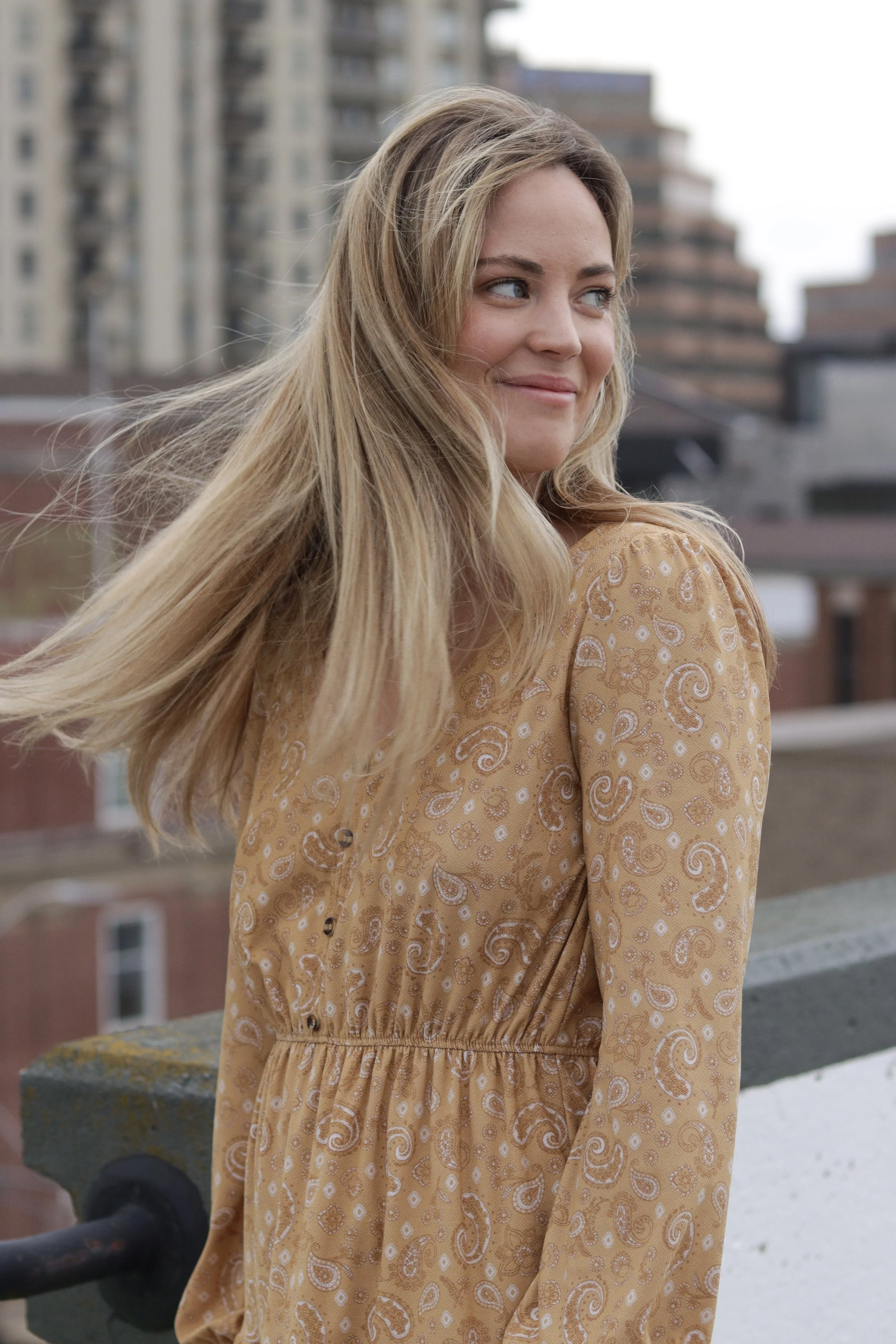 A young woman with long blonde hair wearing a beige dress with a paisley pattern, standing outdoors with a cityscape of tall buildings in the background.