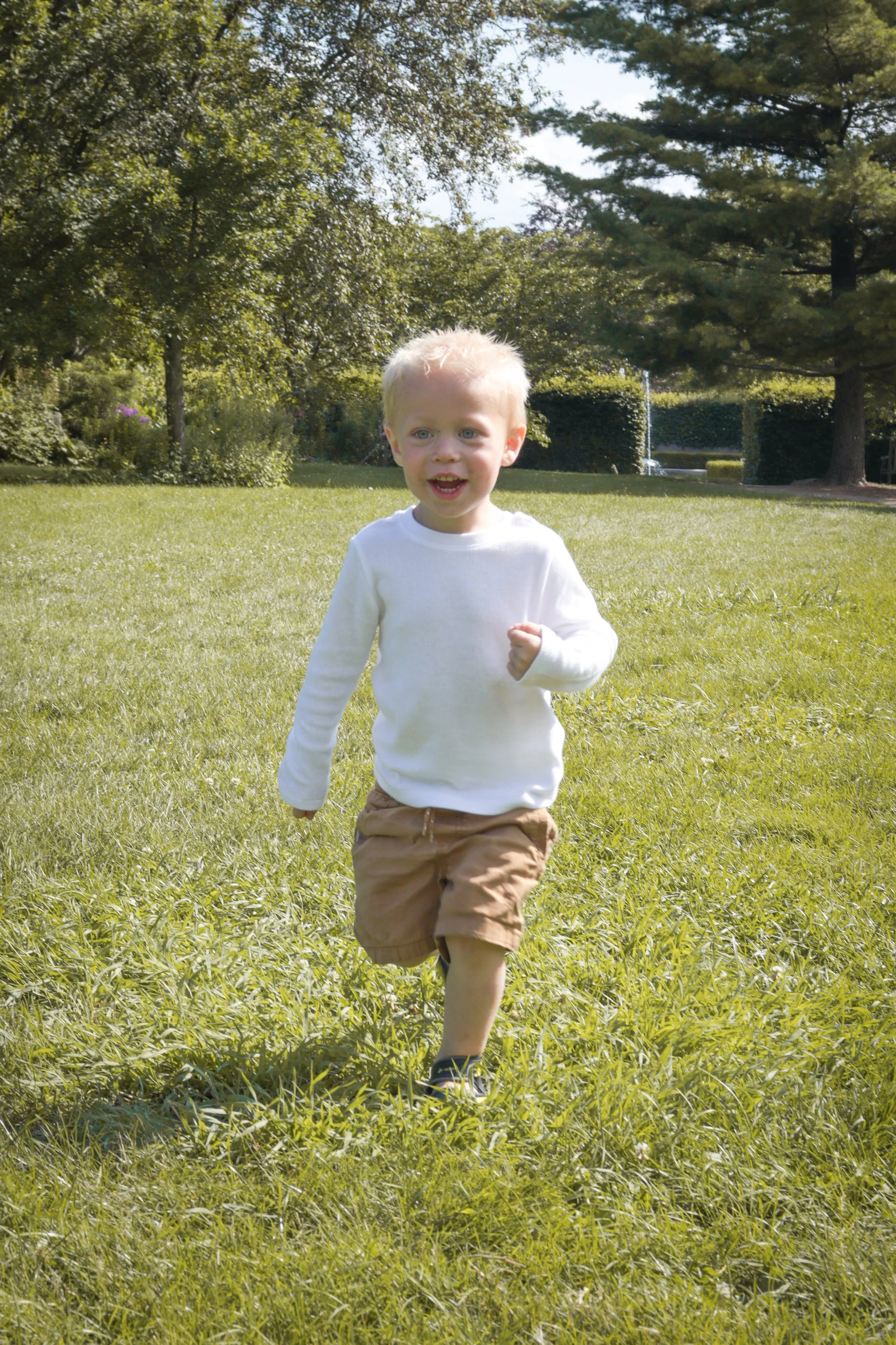 A young boy with blonde hair running across a grassy field on a sunny day.