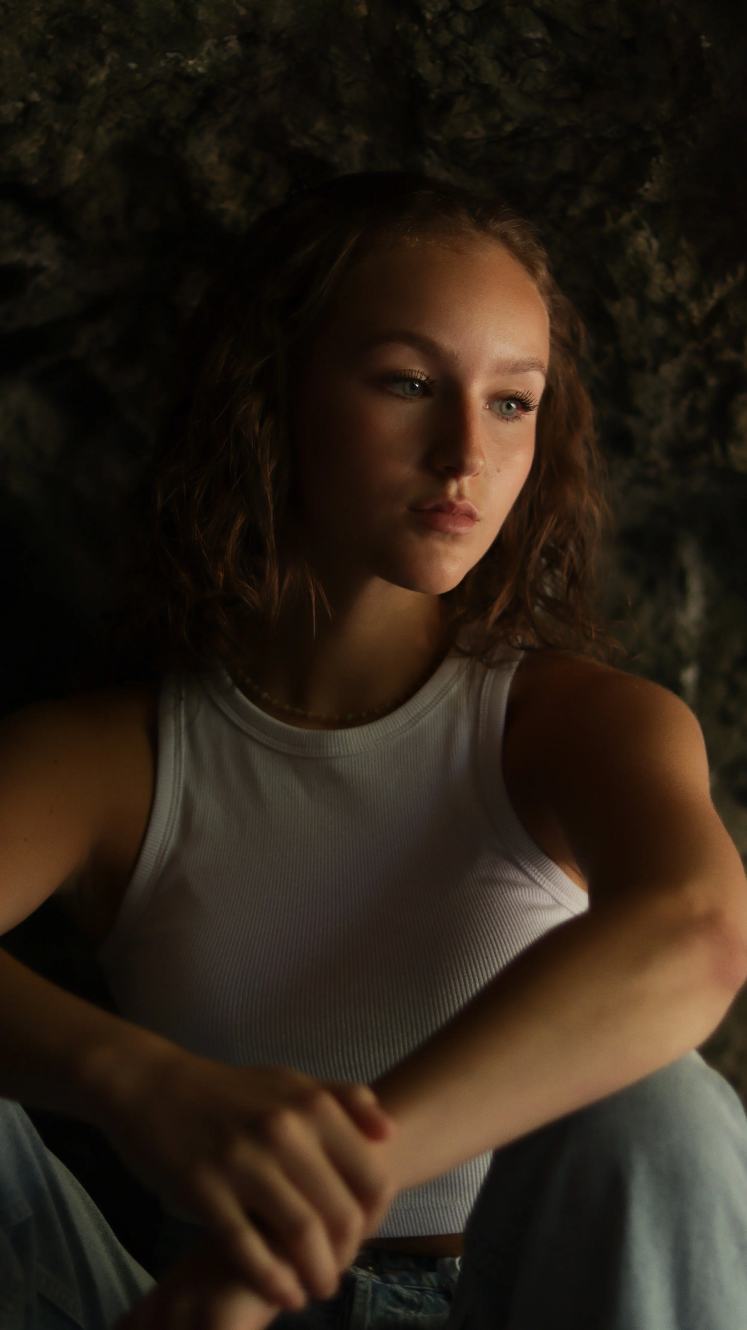 A young woman with light skin and long wavy brown hair, wearing a white sleeveless top, is sitting against a dark, rocky background, looking pensively to her left.