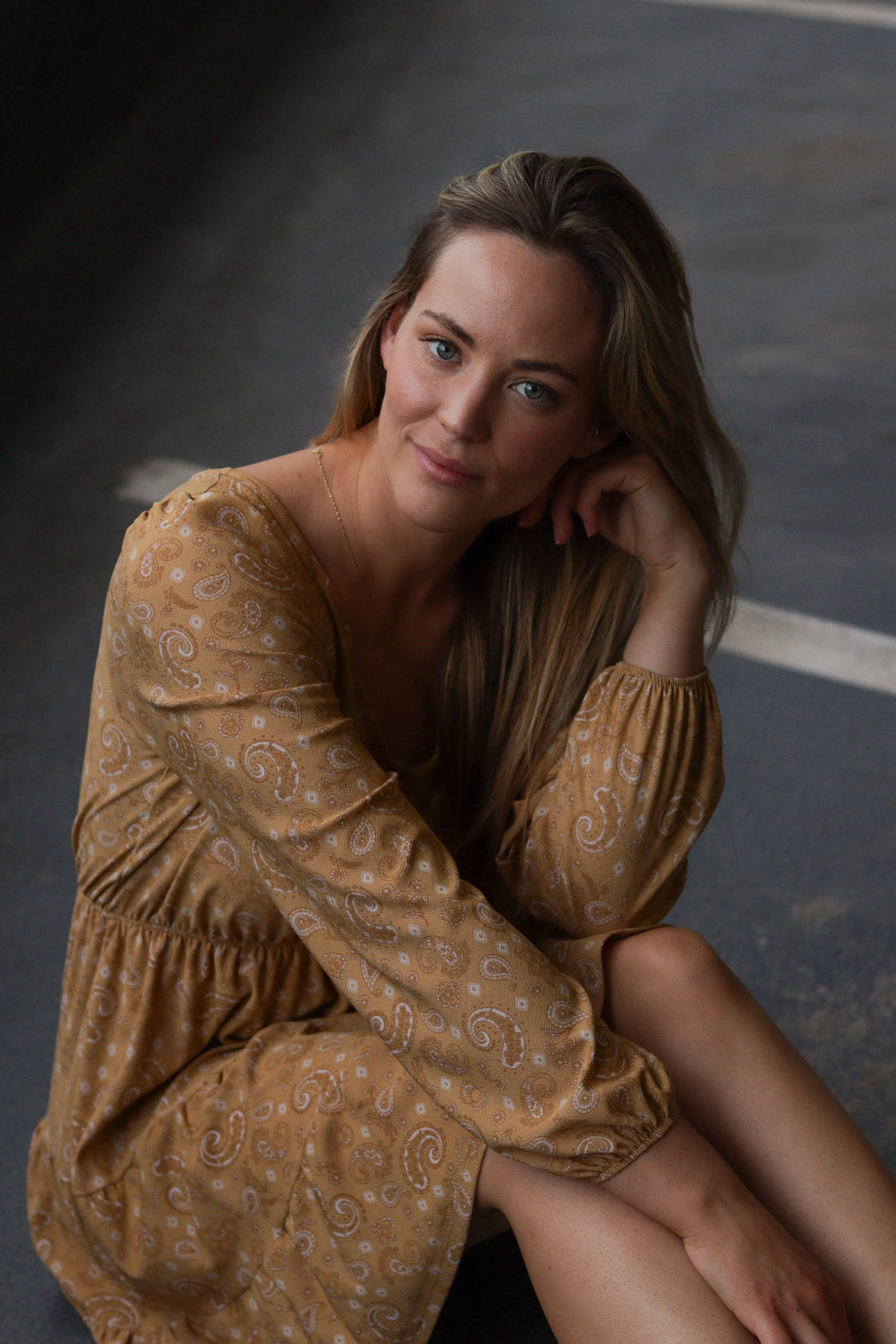 A young woman with long blonde hair, wearing a brown patterned dress, sitting on the ground, looking at the camera with a gentle smile.