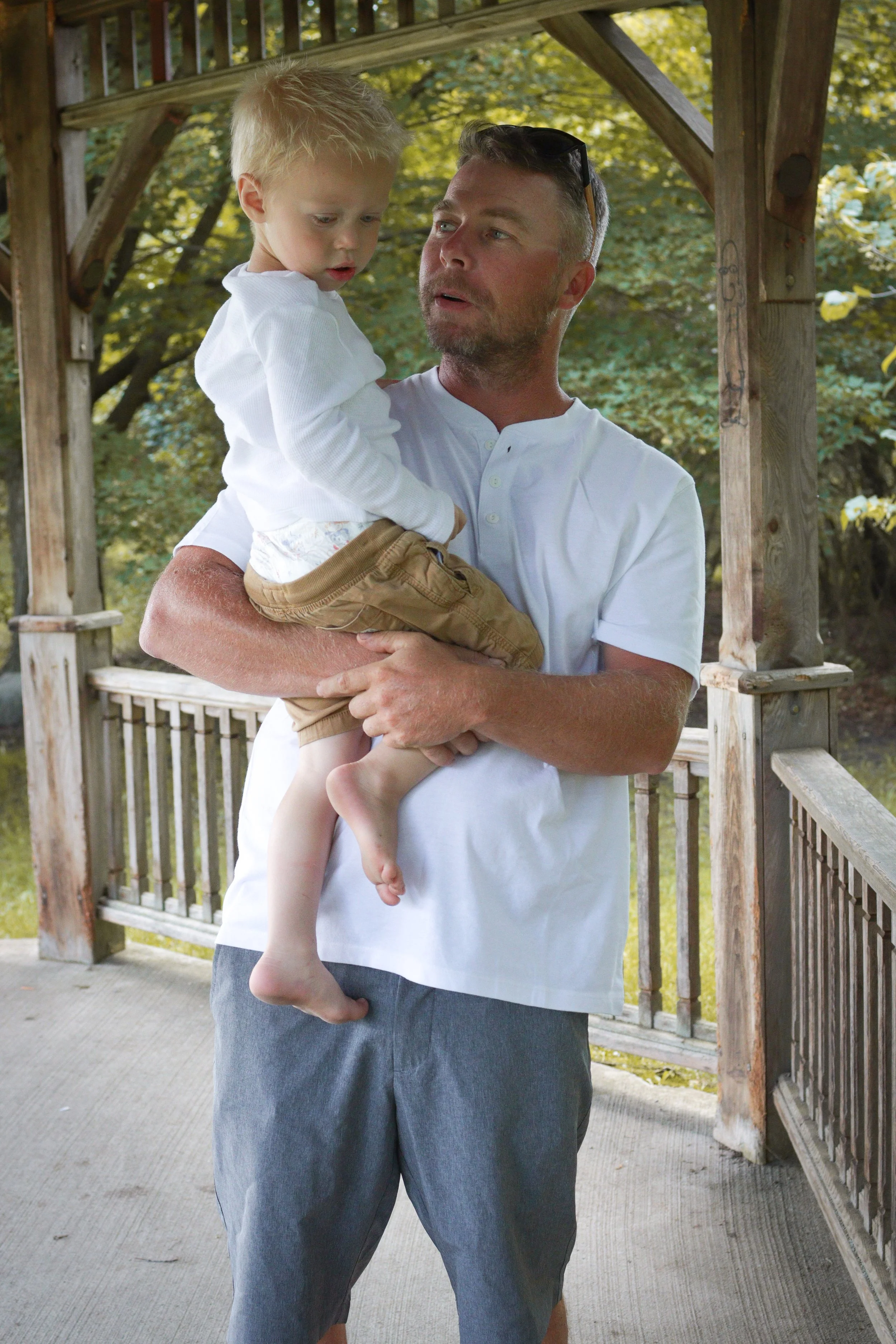 A man holding a young boy in his arms outdoors on a wooden porch, with trees in the background.