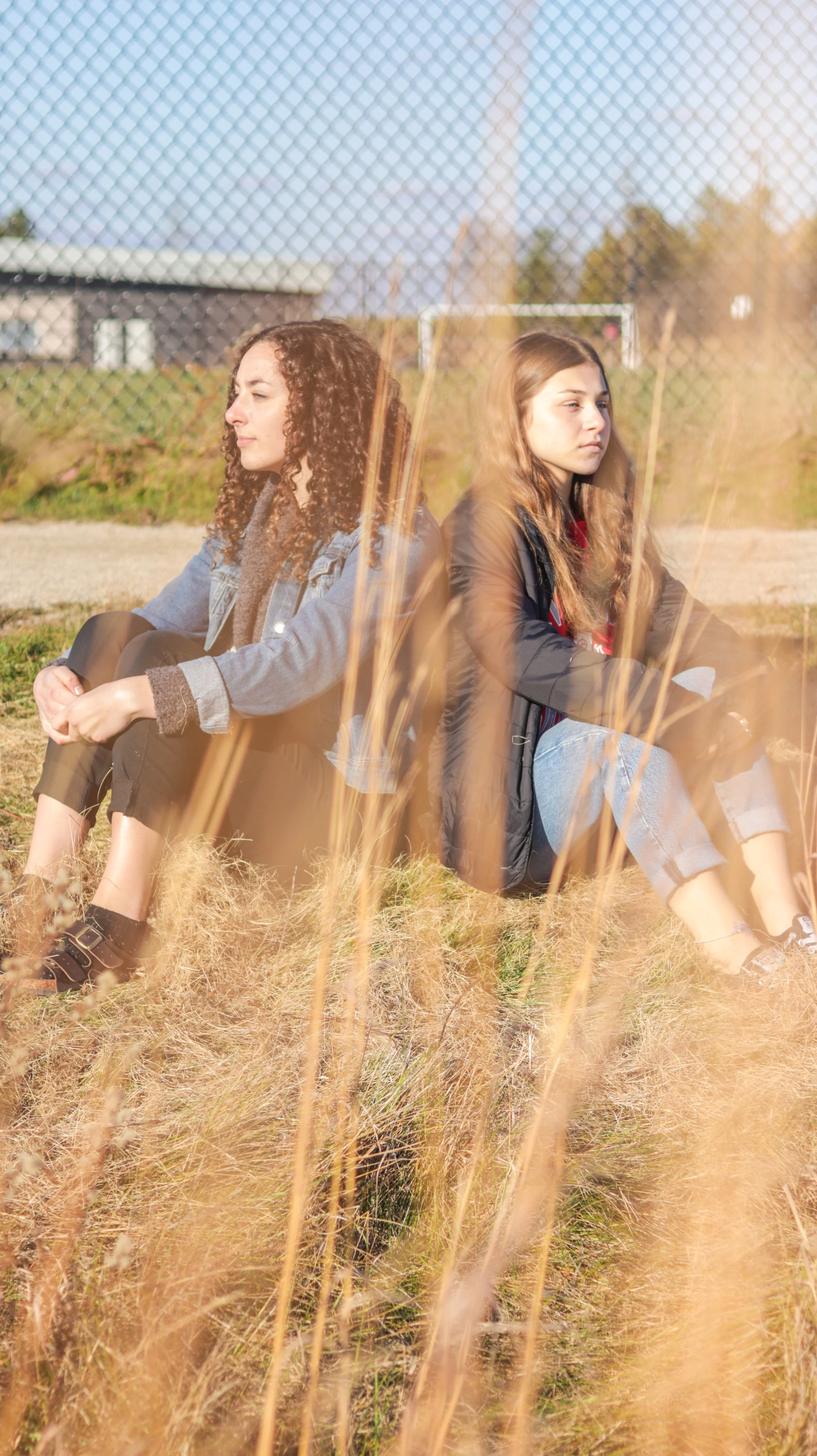 Two young women sitting outdoors on a grassy field, back to back, with a fence and distant trees in the background, during daytime.