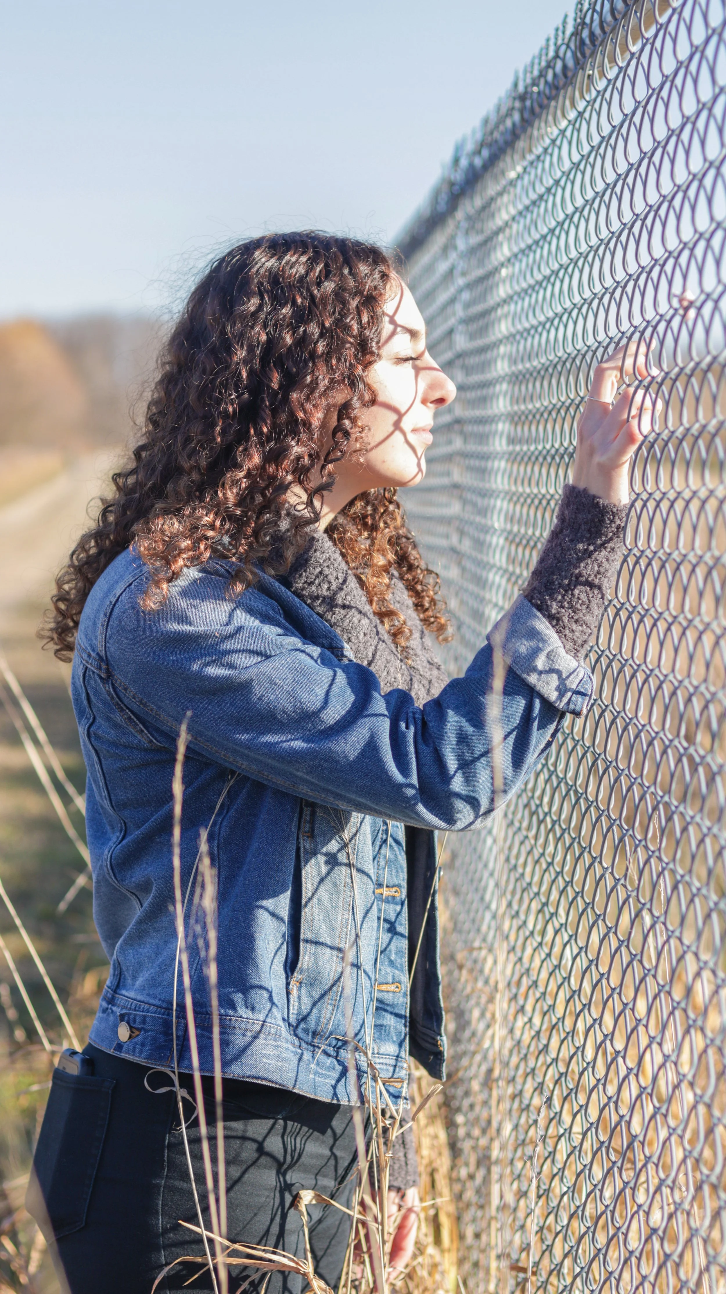 A woman with curly hair, wearing a denim jacket, stands next to a chain-link fence, touching it with her hand, and looking through it with her eyes closed.