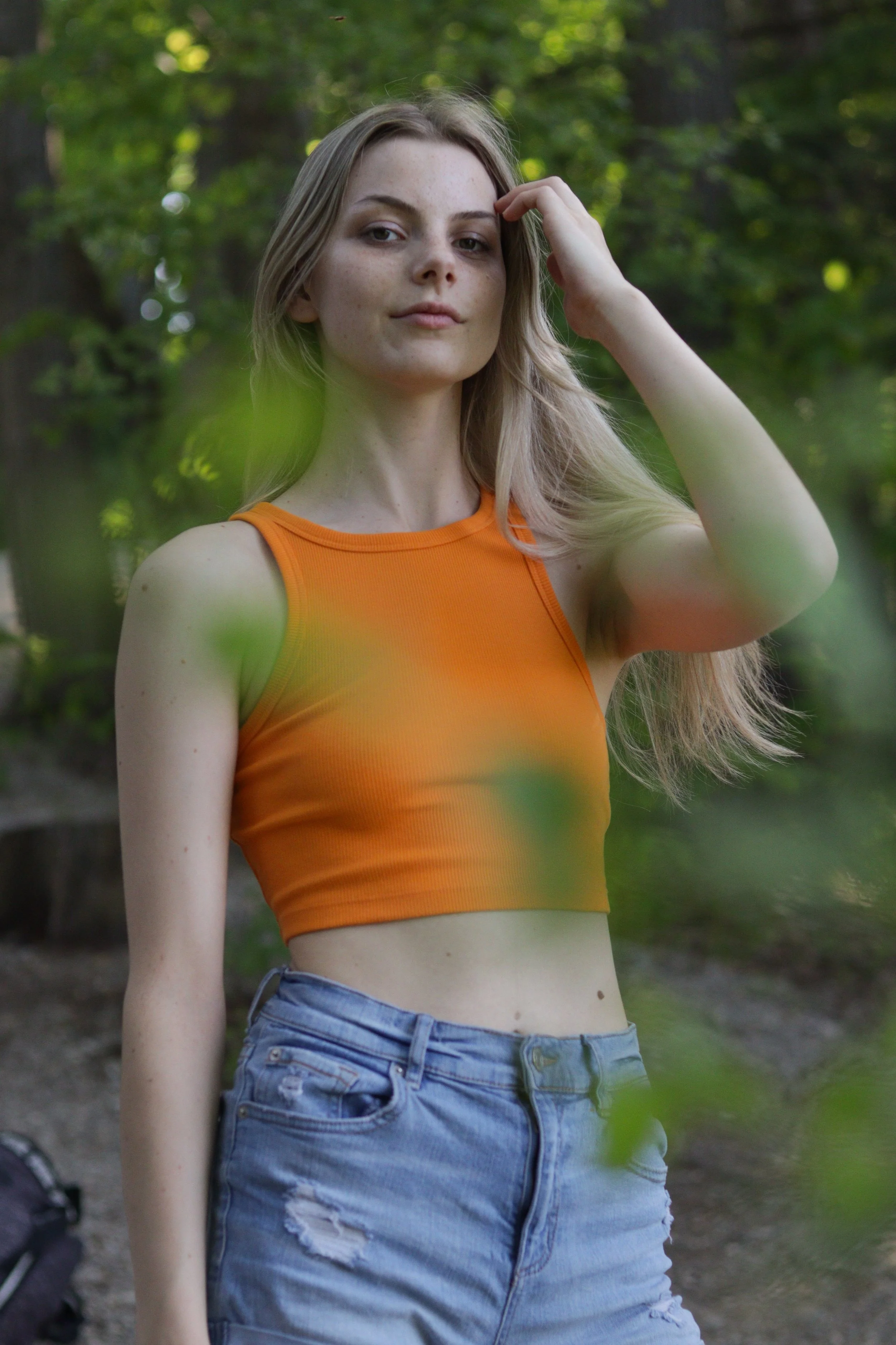 A young woman with long blonde hair wearing an orange sleeveless crop top and distressed denim shorts standing outdoors in a green, wooded area.