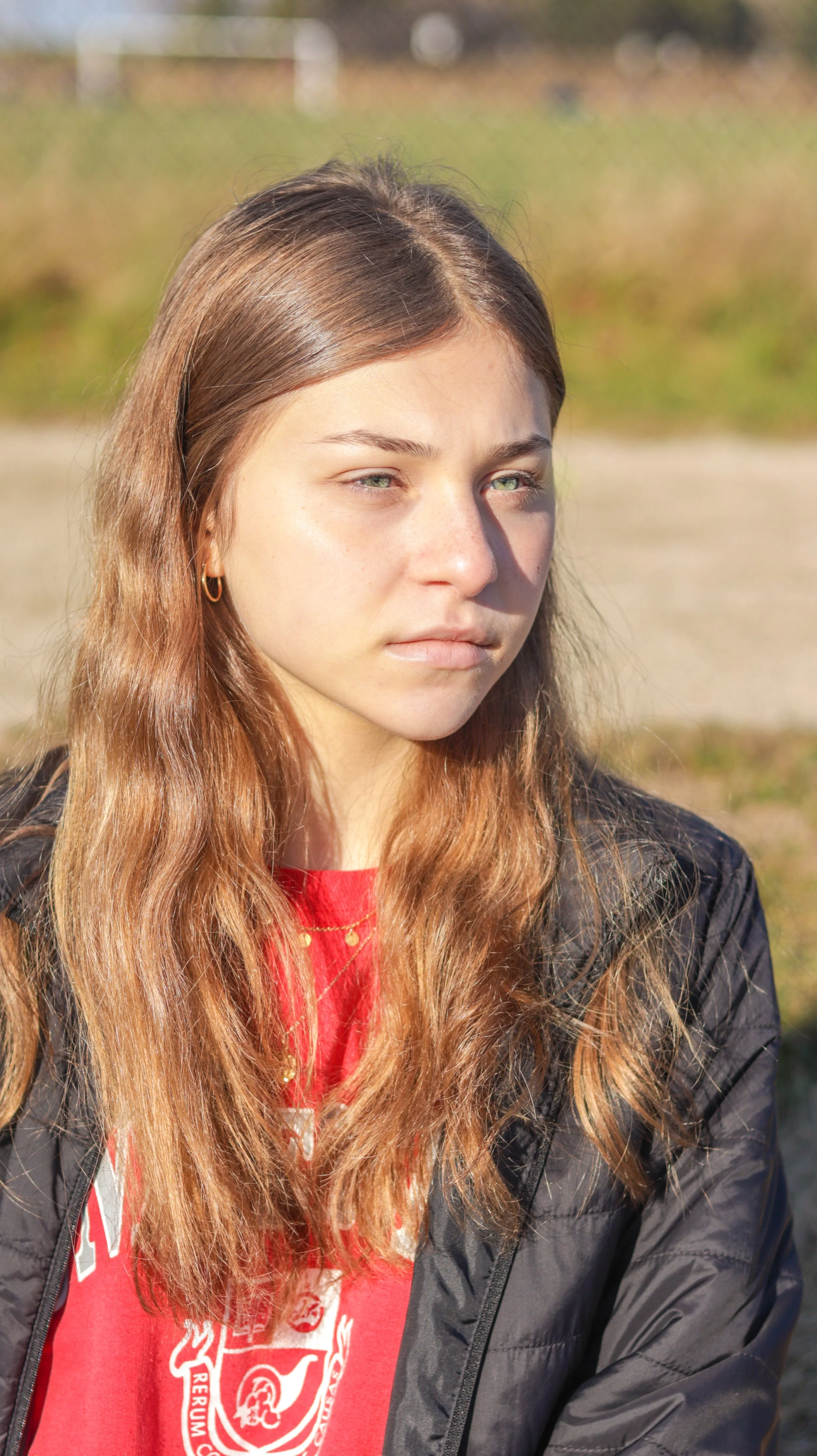 A young woman with light brown hair, wearing a red shirt with a white logo and a black jacket, stands outdoors in sunlight with a slightly serious expression.