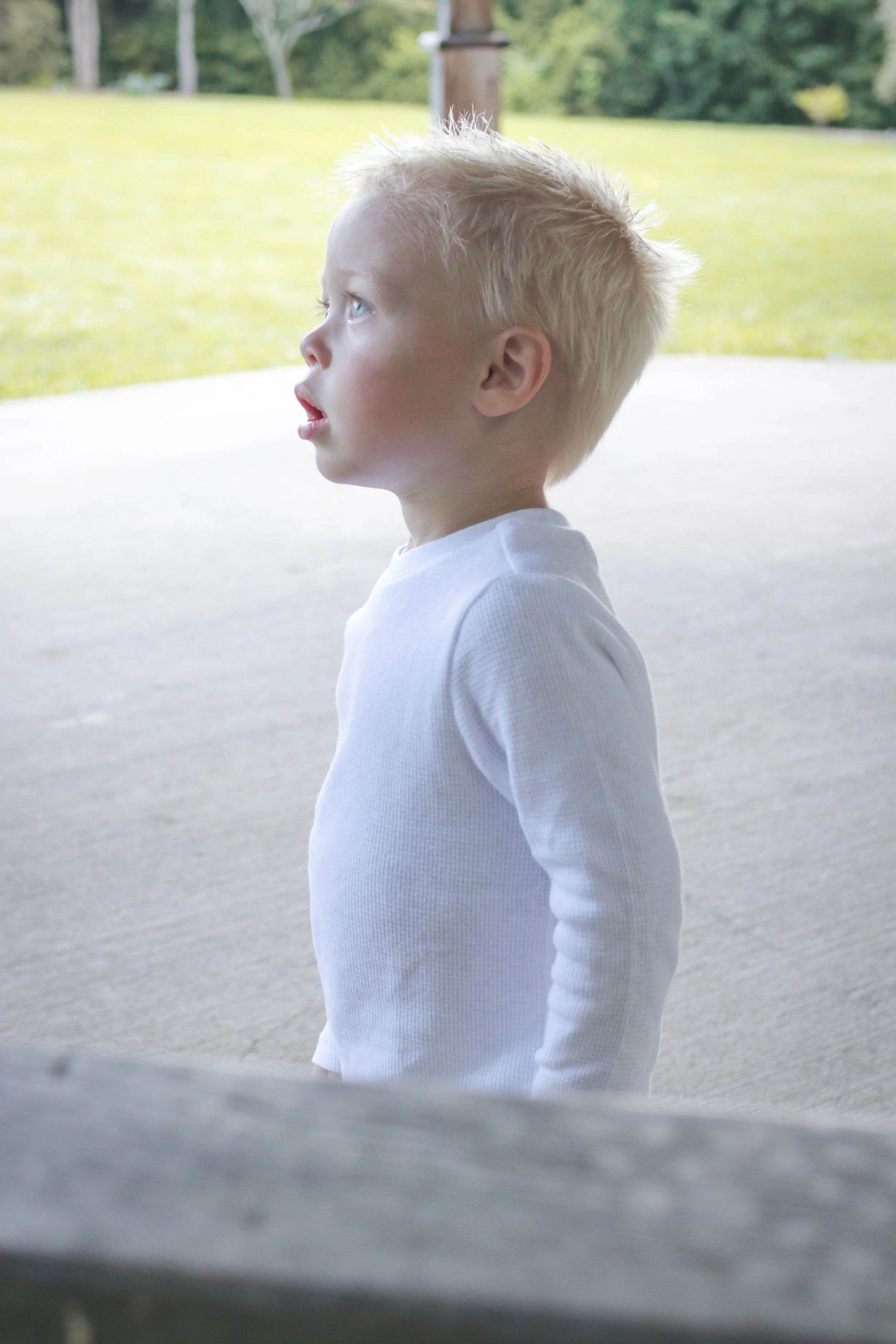 Side profile of a young blonde boy wearing a white long-sleeve shirt, standing outdoors on a concrete surface with grassy area and trees in the background.