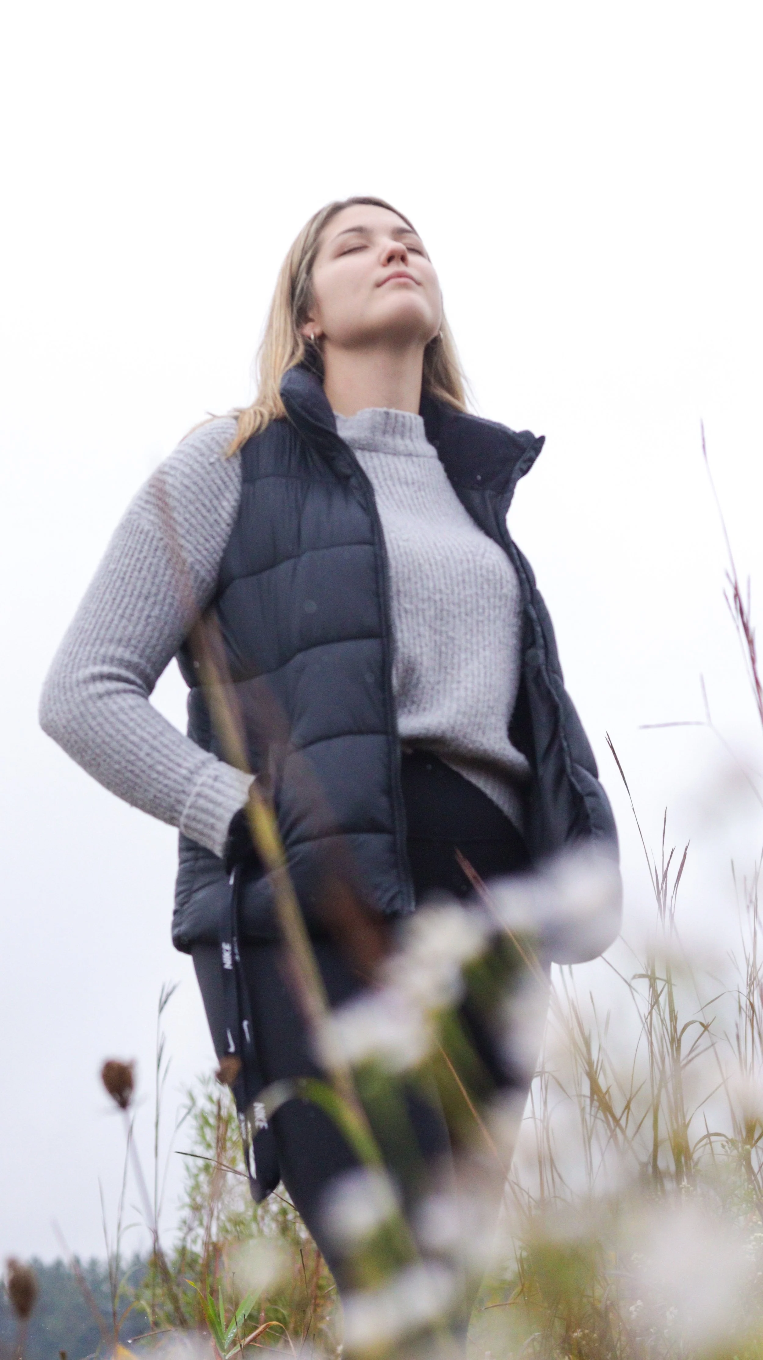 Woman standing outdoors in a field of tall grass, wearing a grey sweater and black vest, with her eyes closed and face tilted upward.