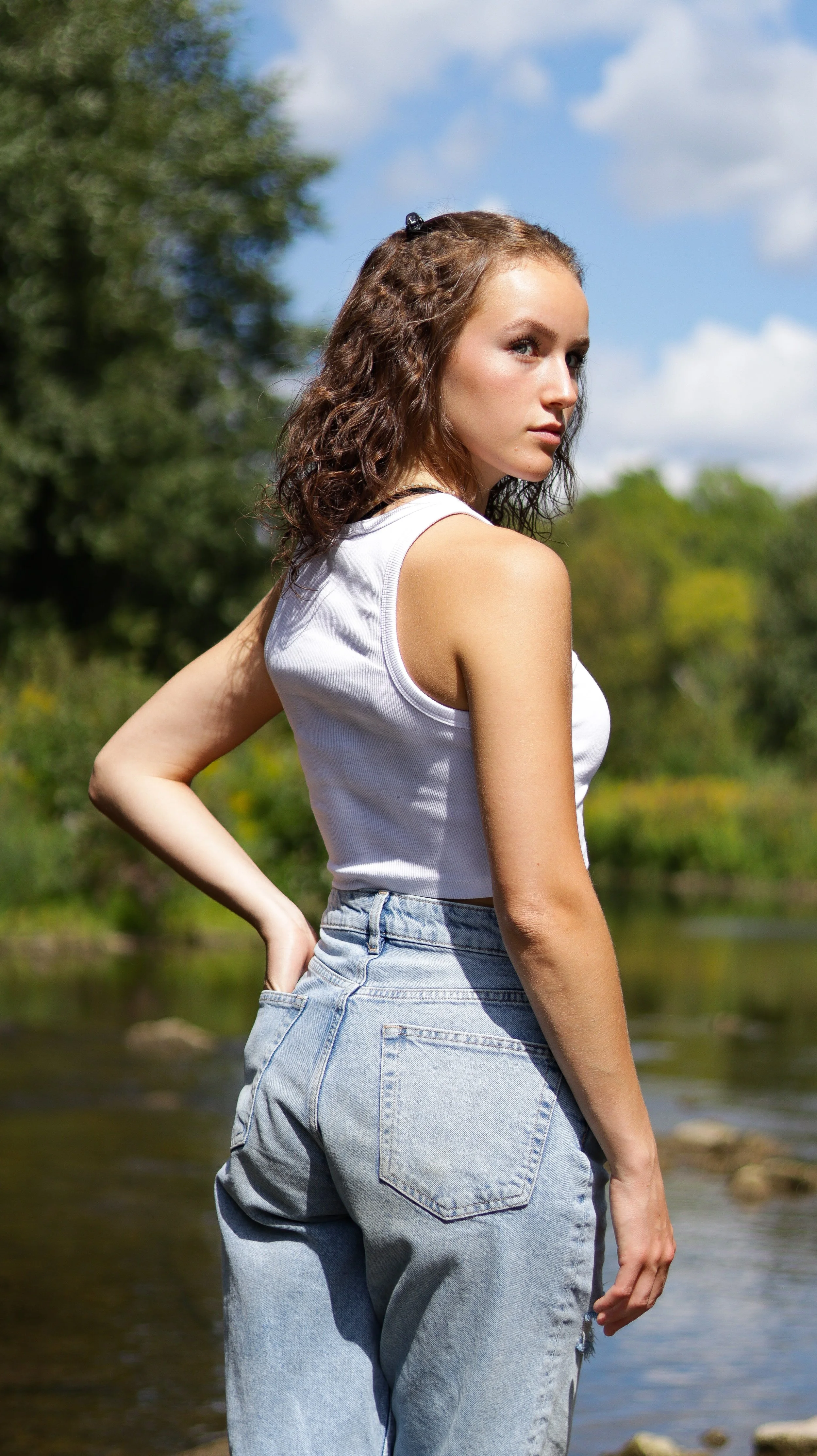 Young woman standing outdoors near a river, wearing a white sleeveless top and high-waisted jeans, with trees and blue sky in the background.