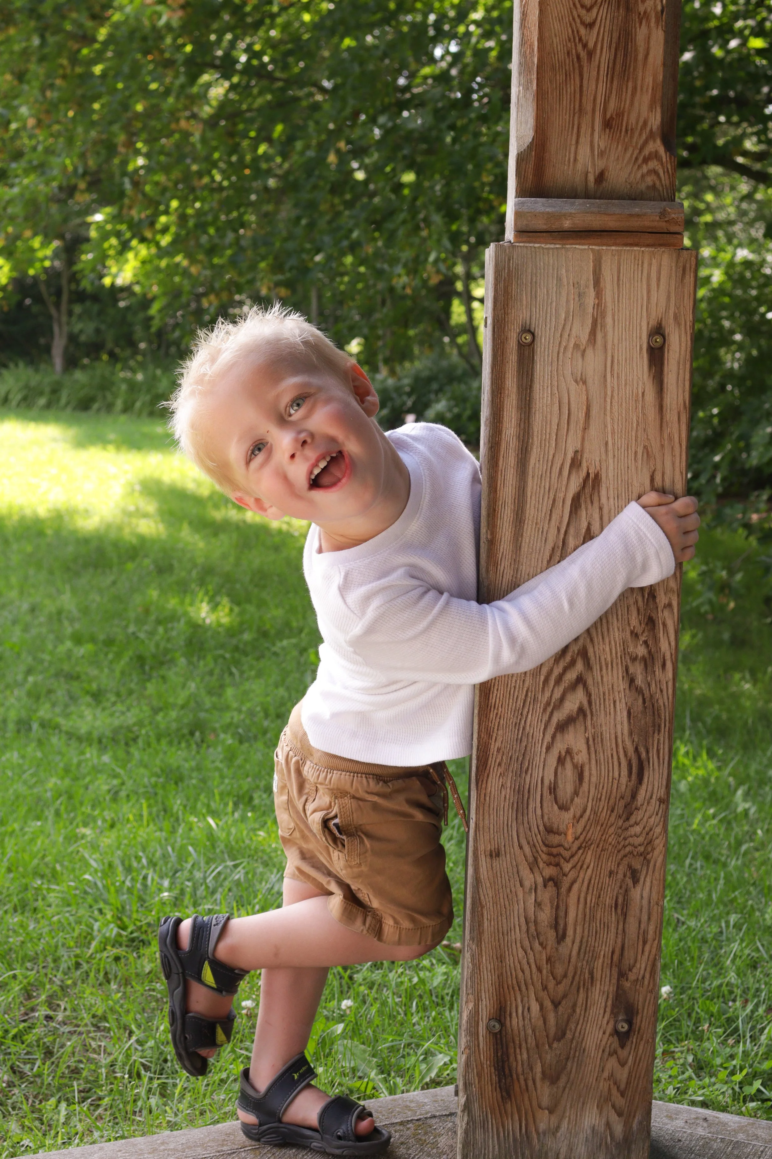 A young boy with blonde hair wearing a white long-sleeve shirt, tan shorts, and sandals, hugging a wooden post outdoors in a grassy area with trees in the background.