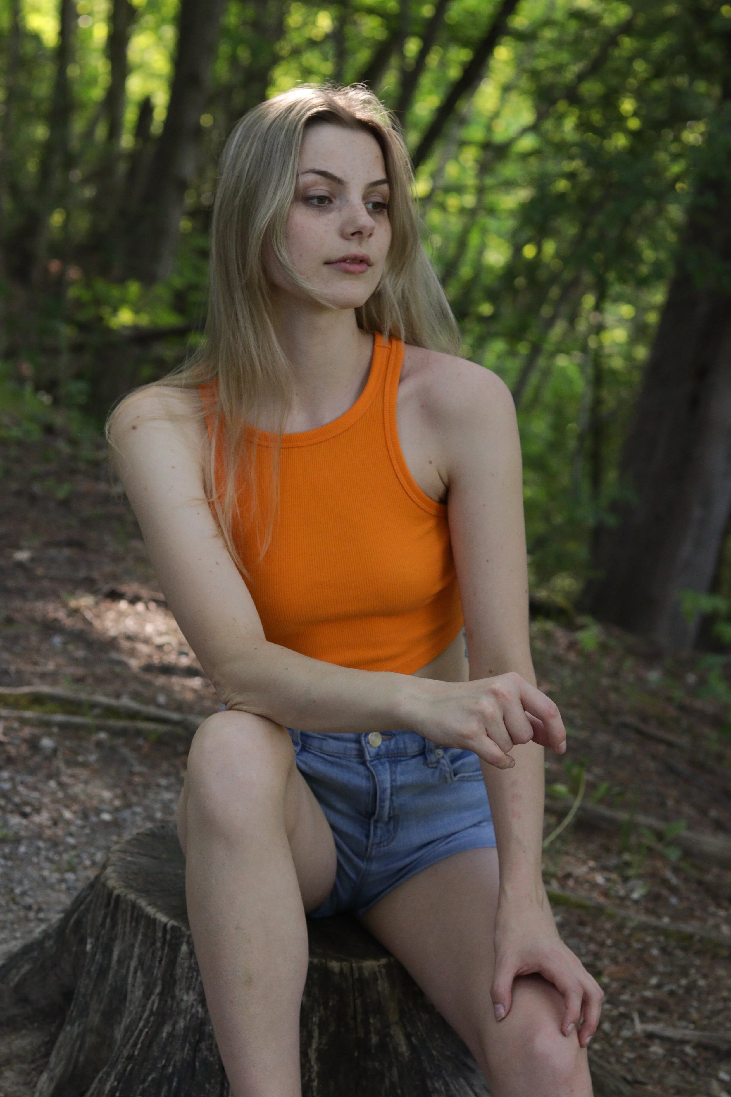 A young woman with long blonde hair, wearing an orange sleeveless top and denim shorts, sitting on a tree stump in a forest.