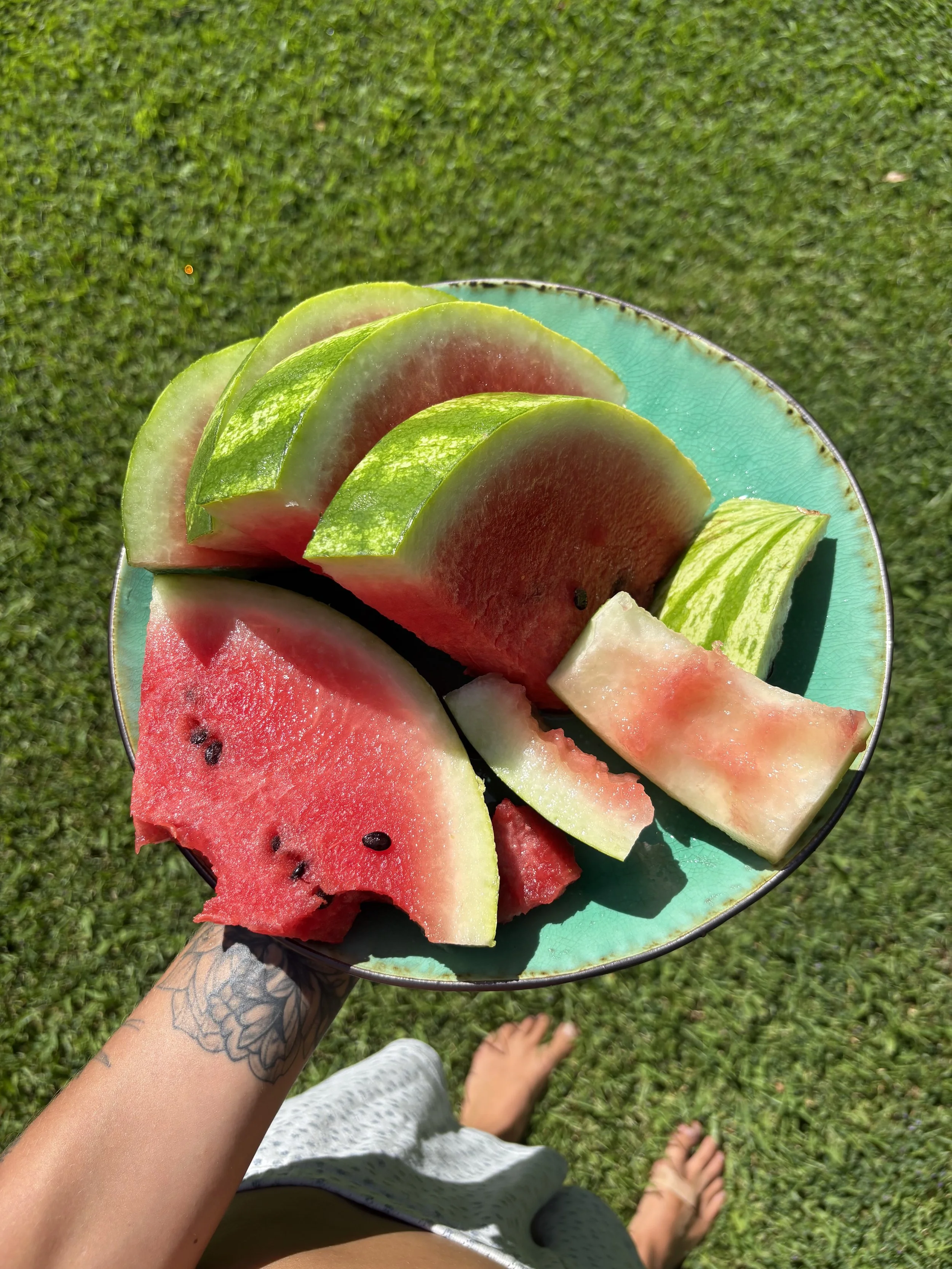 A person holding a turquoise plate with slices of watermelon and cantaloupe, standing on grass with bare feet visible.