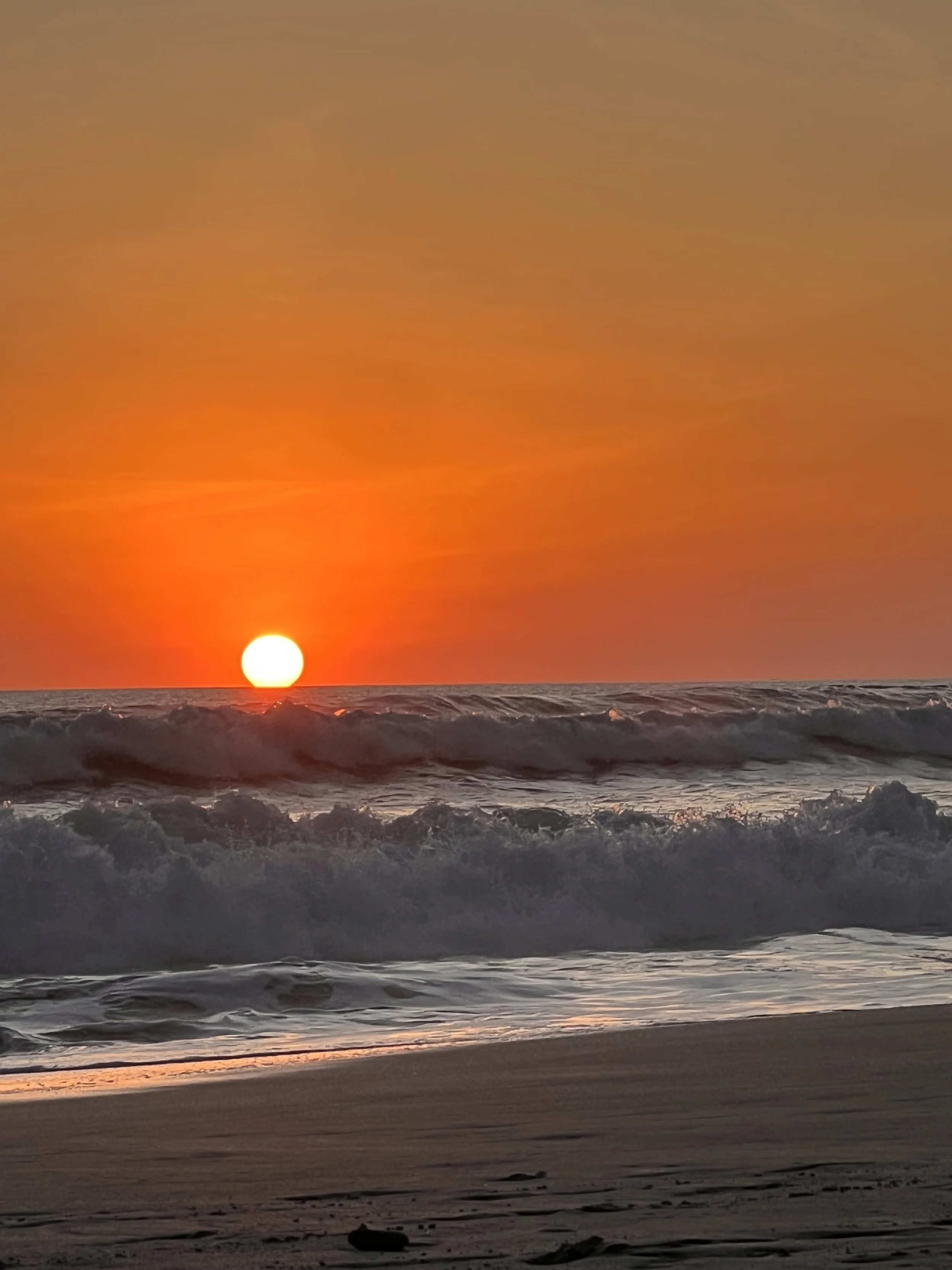 Sun setting over the ocean with waves crashing on the sandy beach.