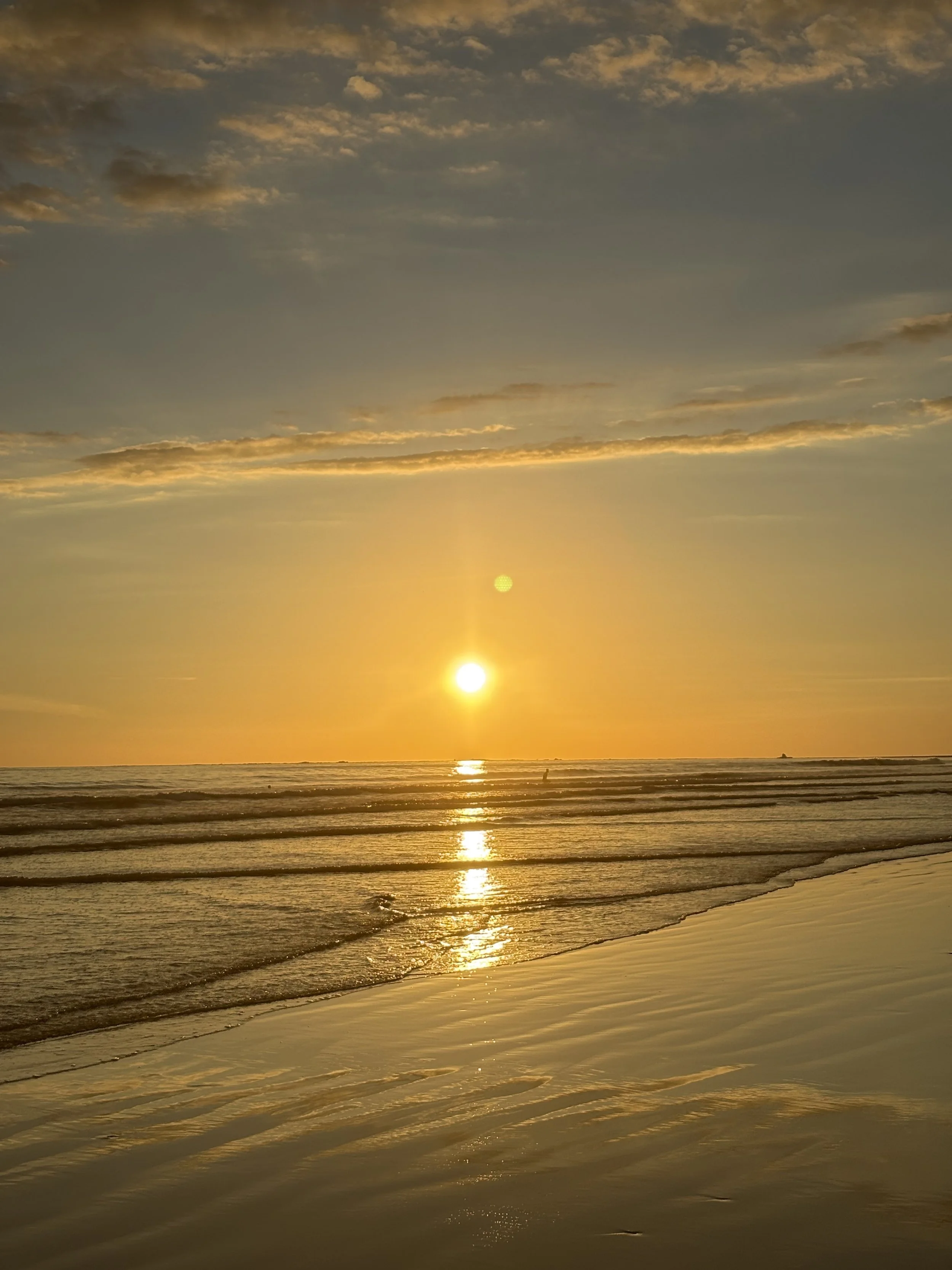 Sunset over the ocean with clouds in the sky and reflections on the water, sandy beach in the foreground.
